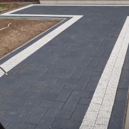 Niko - Ułożona kostka brukowa w odcieniach szarości z białymi akcentami, tworząca ścieżkę prowadzącą do schodów wejściowych. Widoczne donice z kwiatami na schodach.