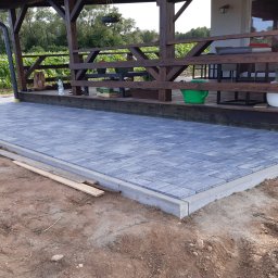 Niko - Ułożona kostka brukowa w odcieniach szarości na tarasie z drewnianą konstrukcją zadaszenia. Widoczne elementy wykończeniowe i przygotowany teren.