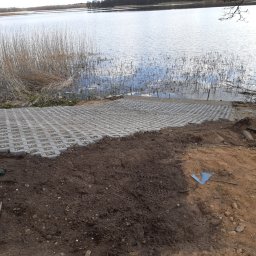 Niko - Ułożona nawierzchnia z ażurowych płyt betonowych nad brzegiem jeziora. Częściowo wyrównany teren, widoczna zagęszczarka. Prace ziemne w toku.