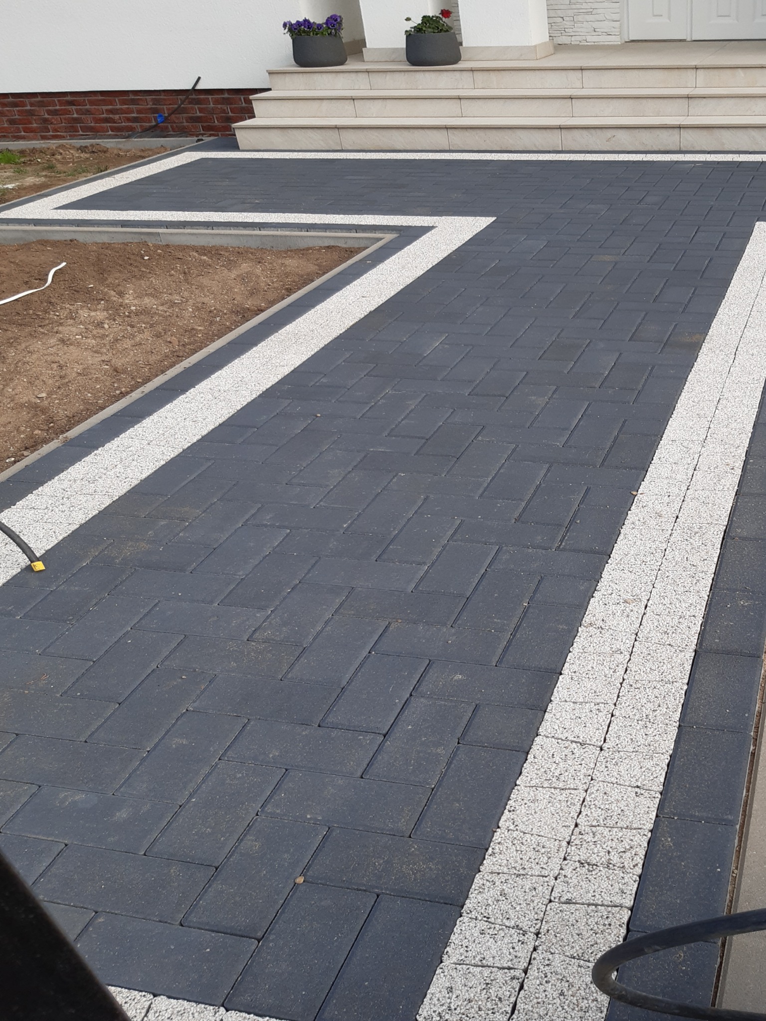 Ułożona kostka brukowa w odcieniach szarości z białymi akcentami, tworząca ścieżkę prowadzącą do schodów wejściowych. Widoczne donice z kwiatami na schodach.
