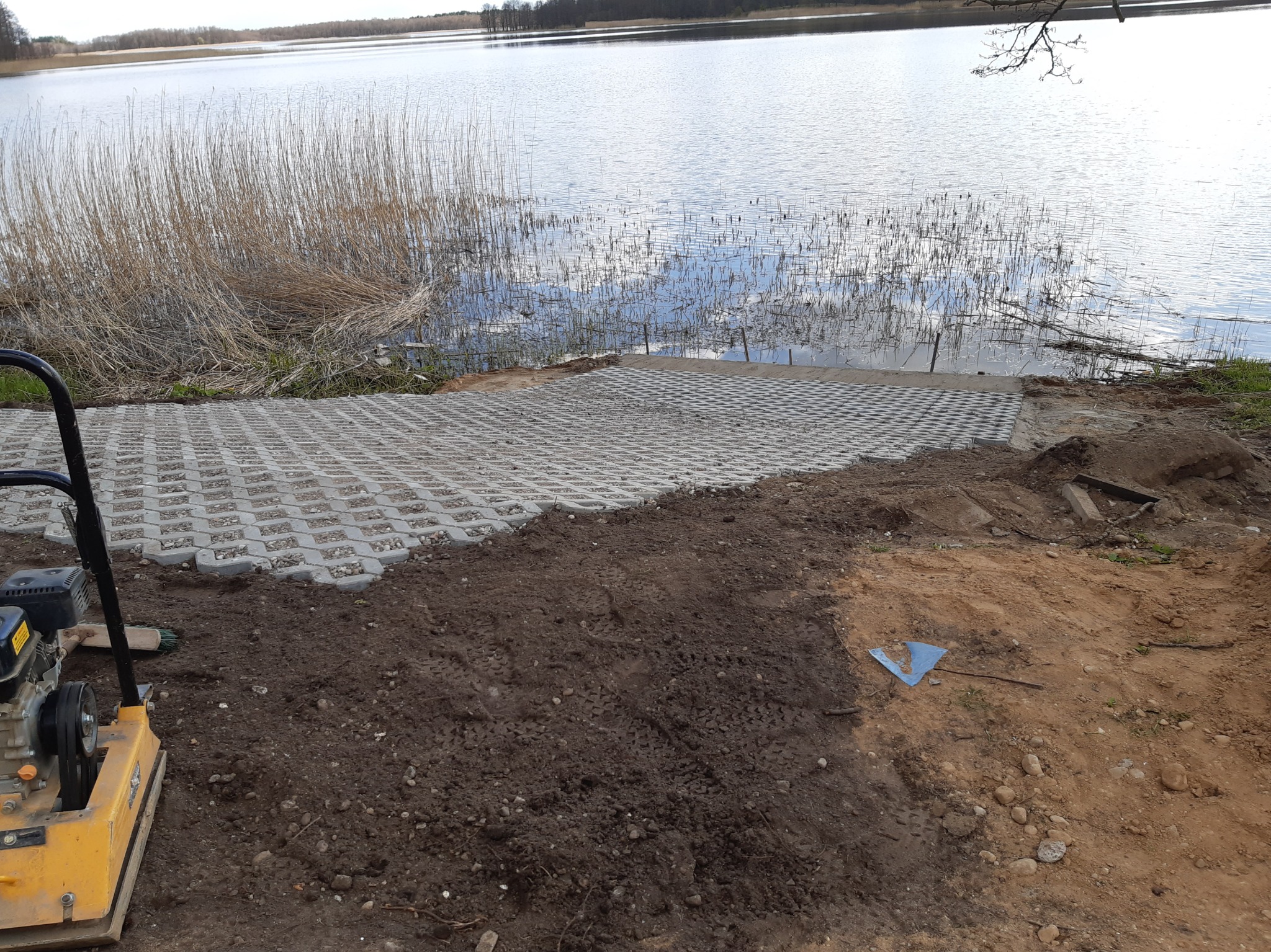 Ułożona nawierzchnia z ażurowych płyt betonowych nad brzegiem jeziora. Częściowo wyrównany teren, widoczna zagęszczarka. Prace ziemne w toku.