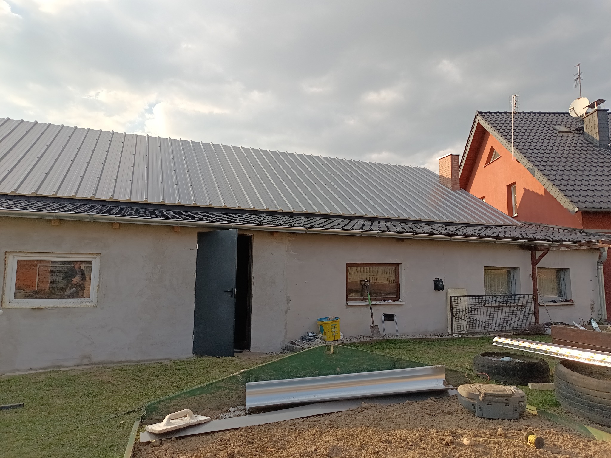 Szary budynek z nowym, błyszczącym dachem z blachy trapezowej. Otwarta brama garażowa, okna z widocznymi narzędziami wewnątrz. W tle dom z czerwoną elewacją i ciemnym dachem.