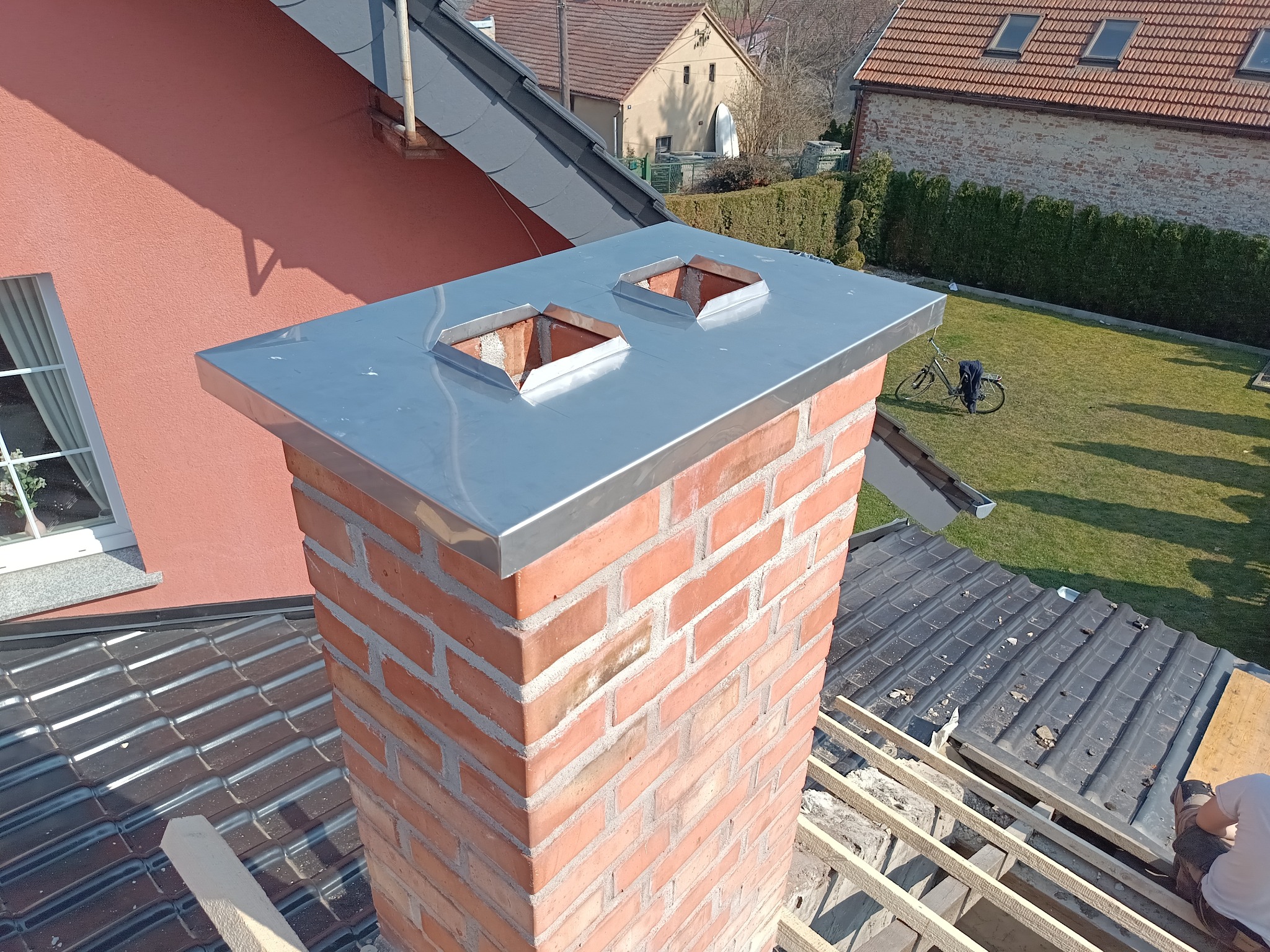 Ceglany komin z nową, błyszczącą, metalową obudową na dachu z częściowo zdjętą dachówką. W tle domy i zieleń.