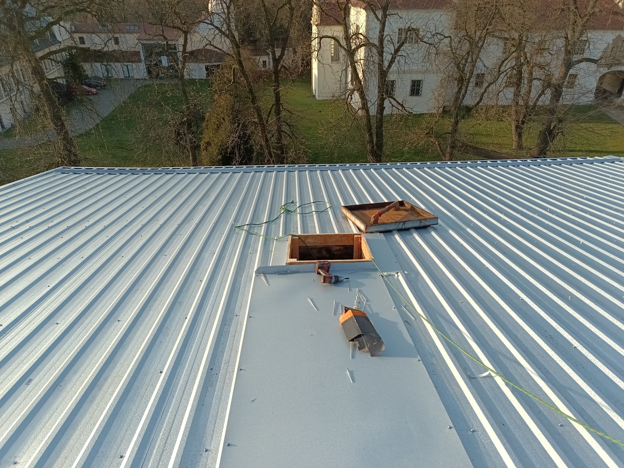 Montaż okna dachowego na blaszanym dachu z widocznymi narzędziami i elementami konstrukcyjnymi. W tle budynki i drzewa.