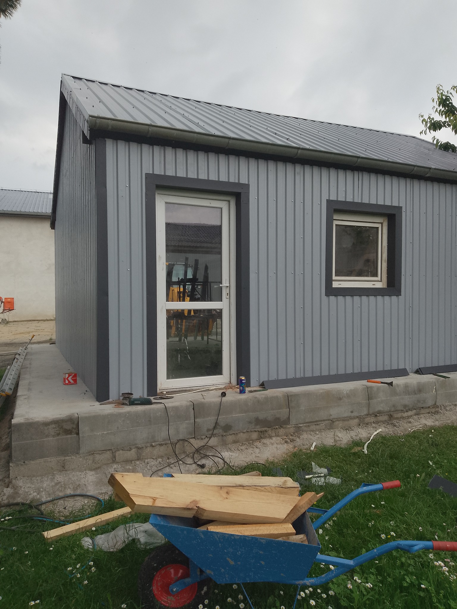 Szary blaszany budynek z oknem i drzwiami, widoczne narzędzia i taczka z drewnem, sugerujące prace wykończeniowe dachu i elewacji. Detal: ciemne obramowania okien i drzwi.