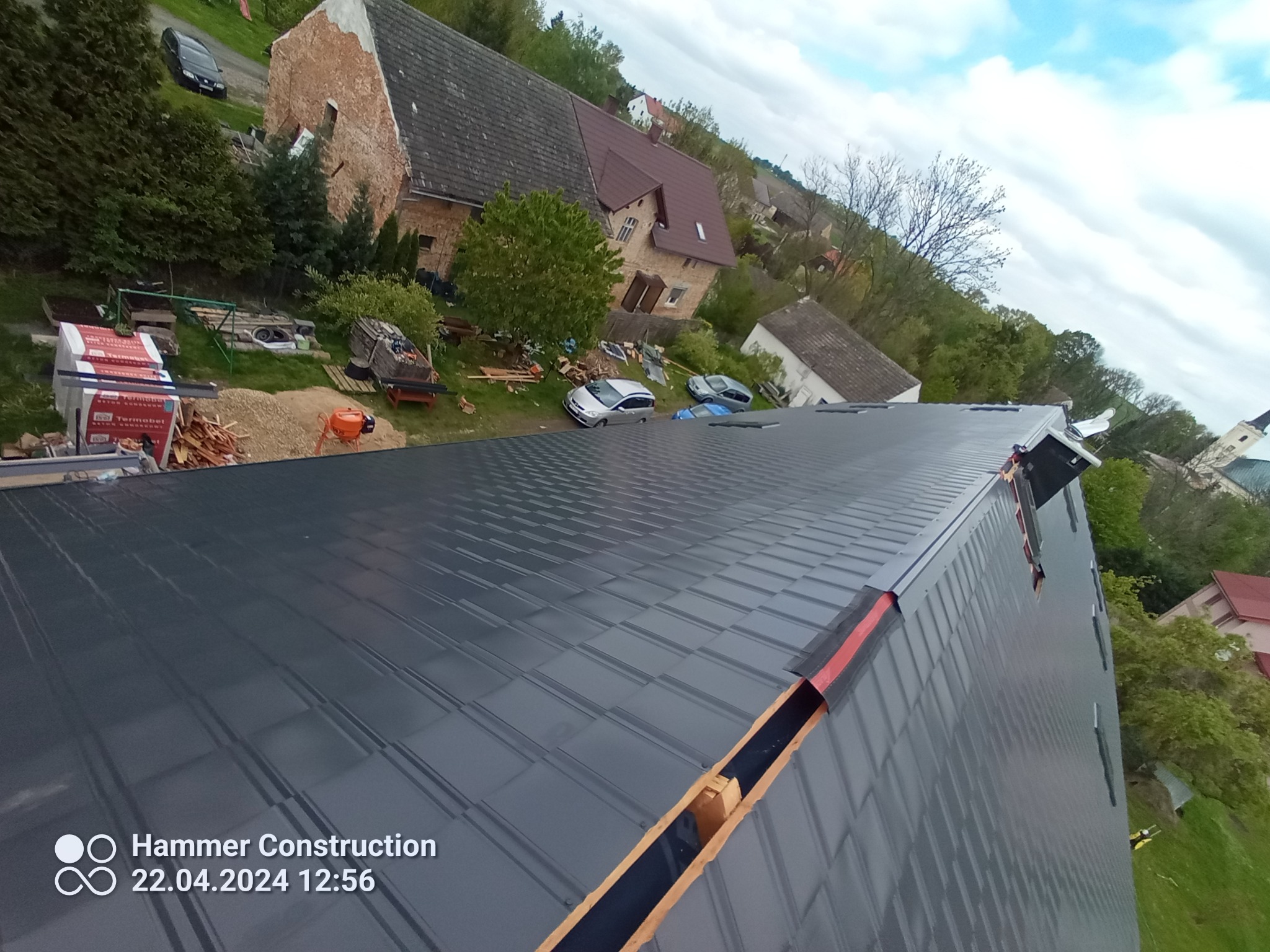 Nowy, grafitowy dach z blachy trapezowej w Głogówku, z widocznymi elementami montażowymi i otoczeniem budynków mieszkalnych. Ujęcie z góry.