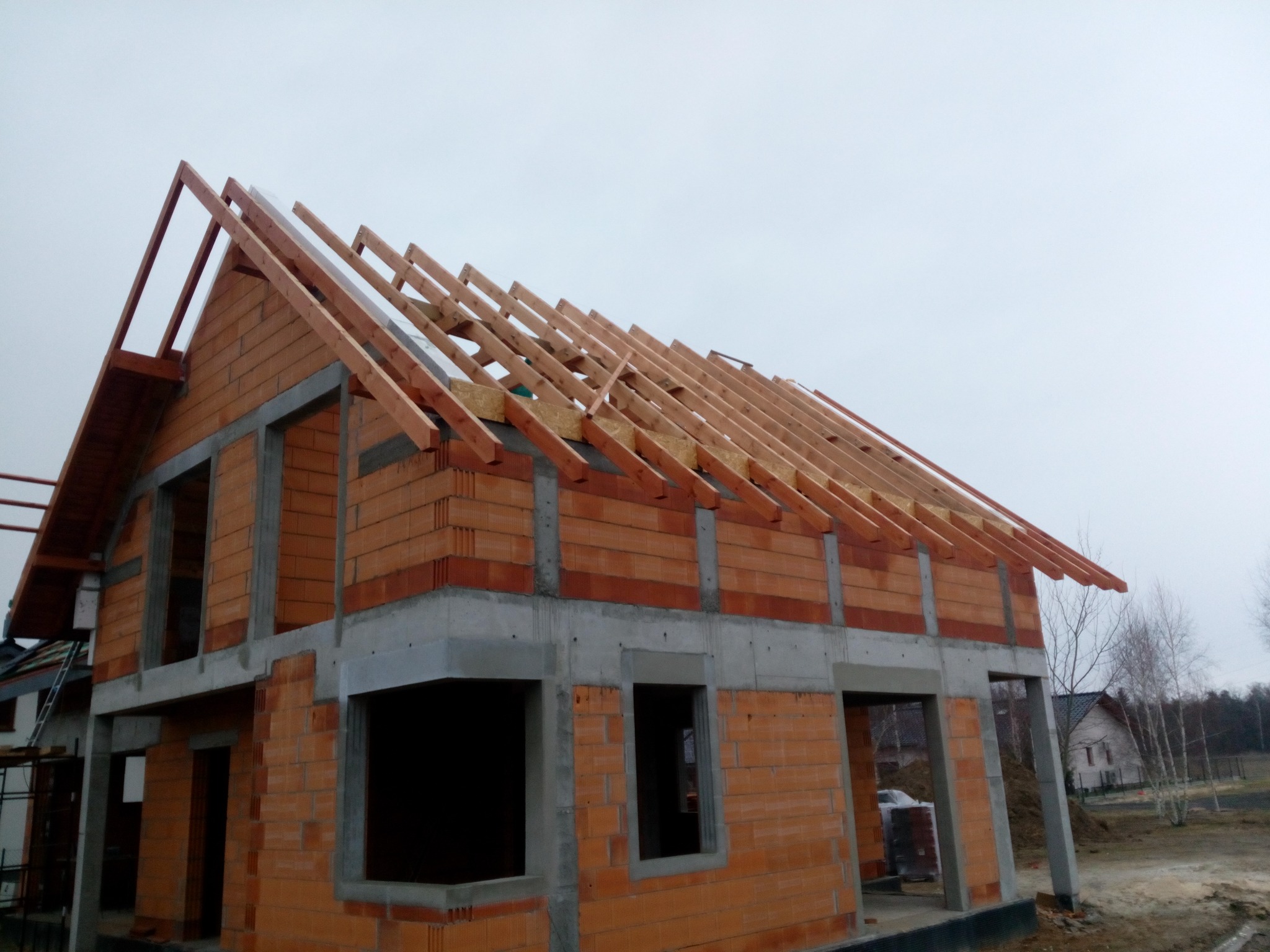 Surowy stan domu z czerwonej cegły z widoczną konstrukcją drewnianą dachu na tle pochmurnego nieba. Widoczne elementy betonowe i pustaki.