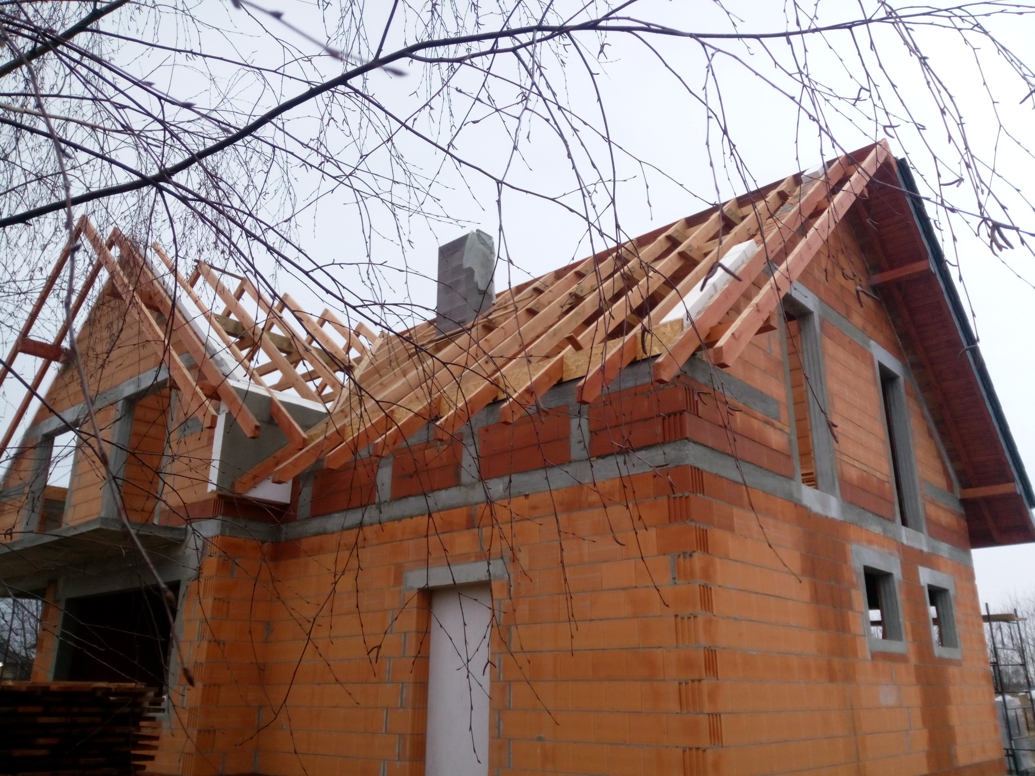 Surowy stan budynku z czerwonej cegły z widoczną konstrukcją dachu z drewna, komin z szarego betonu, na tle pochmurnego nieba i gałęzi drzew.