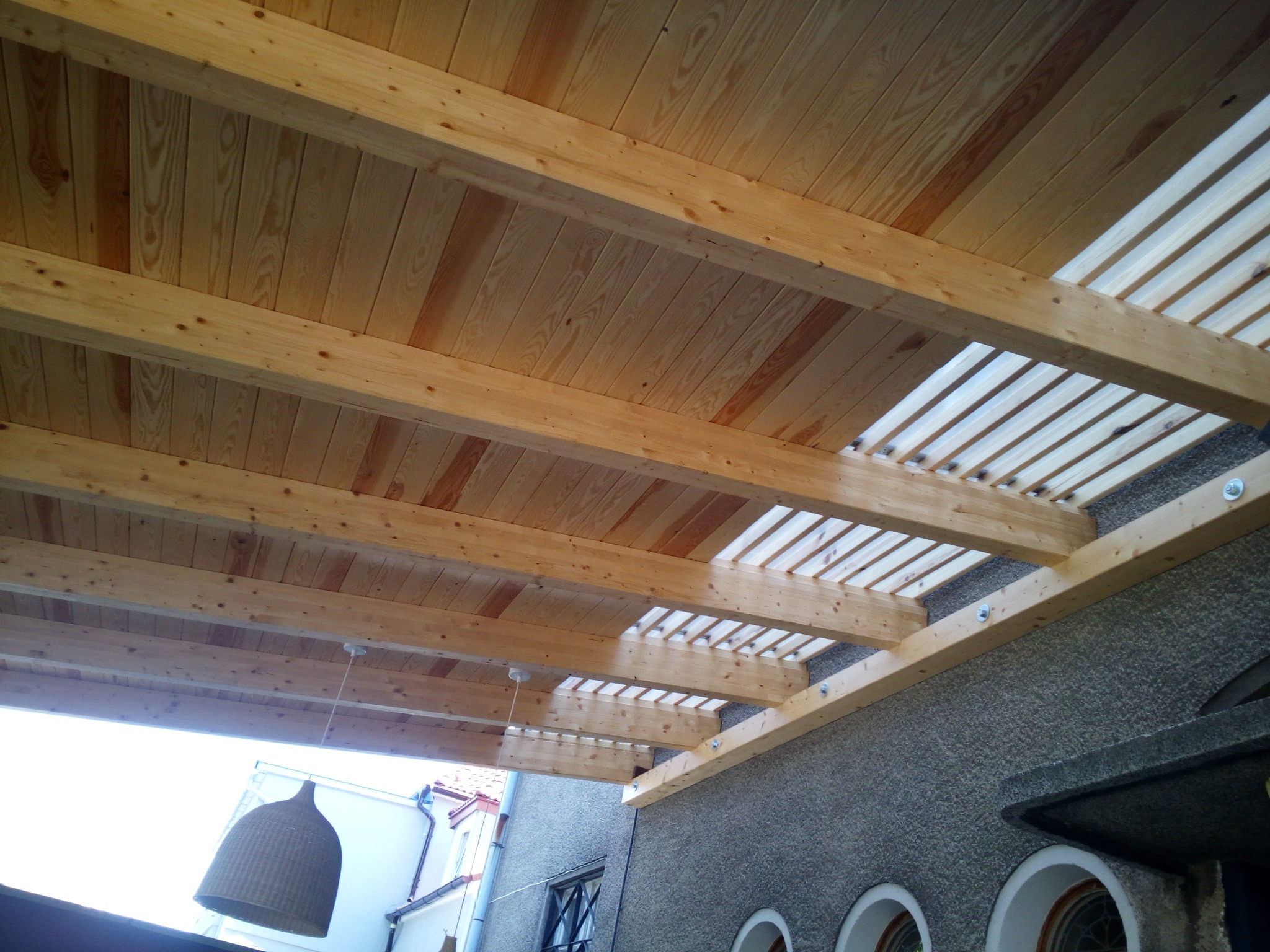 Drewniana konstrukcja z elementami zadaszenia, widok z dołu, z fragmentem elewacji budynku i lampą wiszącą. Jasne drewno, widoczne łączenia śrubowe.