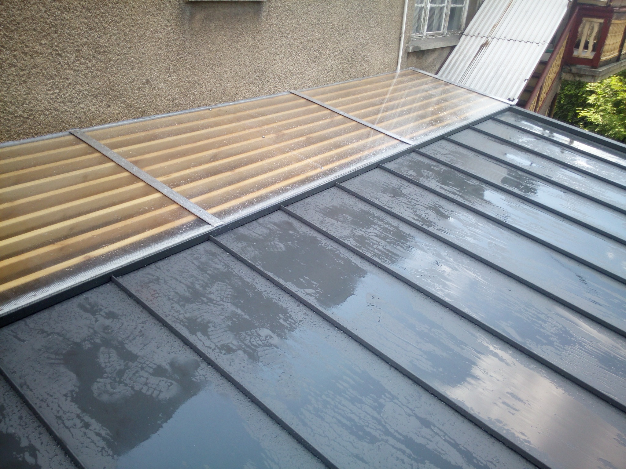 Szary dach z blachy na rąbek stojący, po deszczu, z fragmentem przezroczystego zadaszenia i widoczną elewacją budynku w tle.