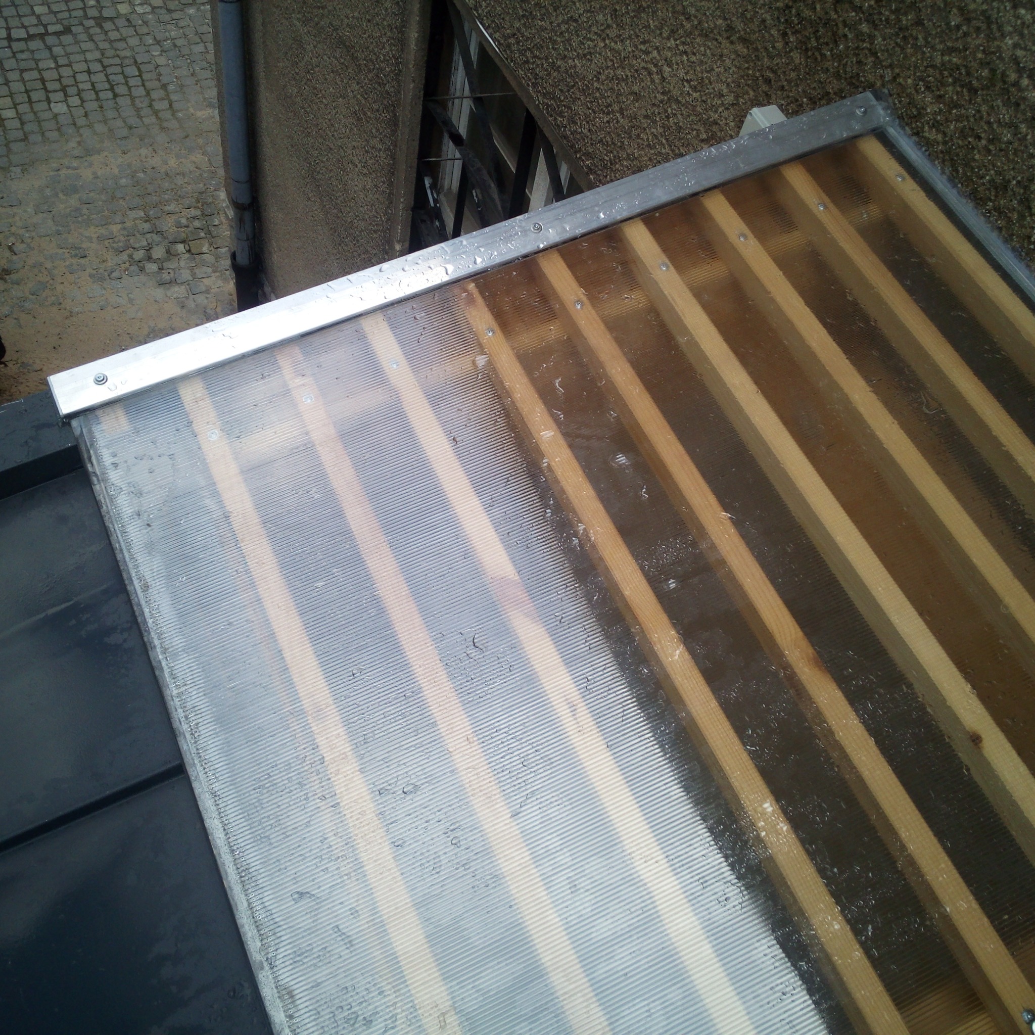 Fragment dachu z przezroczystym poliwęglanem i drewnianą konstrukcją nośną, zabezpieczony metalową listwą. Widoczne krople wody.