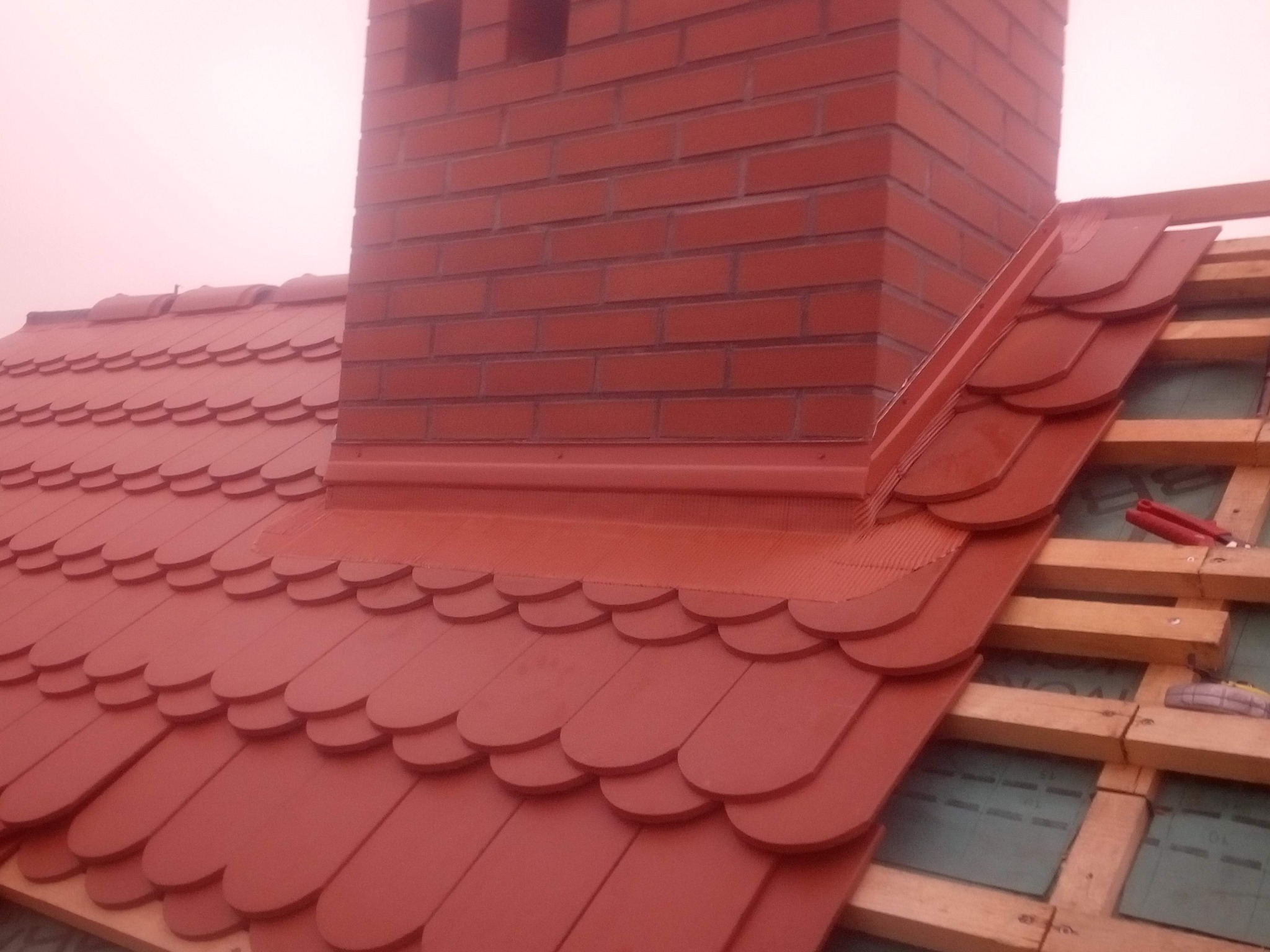 Czerwony dach z dachówki karpiówki i ceglany komin. Widoczne elementy więźby dachowej. Detal wykonania obróbek blacharskich wokół komina.