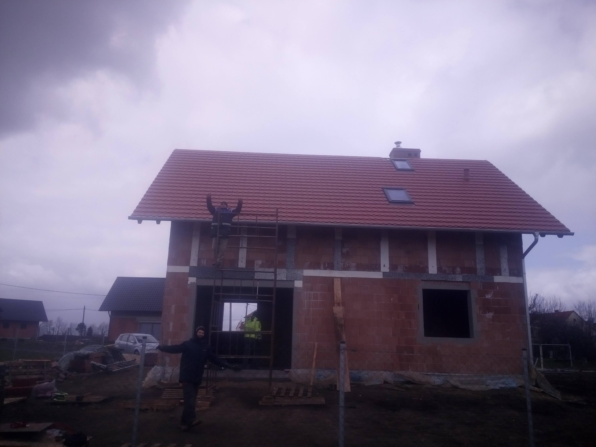 Dom w trakcie budowy z czerwoną dachówką, widoczni dwaj pracownicy na rusztowaniu. Jeden z nich unosi ręce w geście triumfu. Pochmurne niebo w tle.