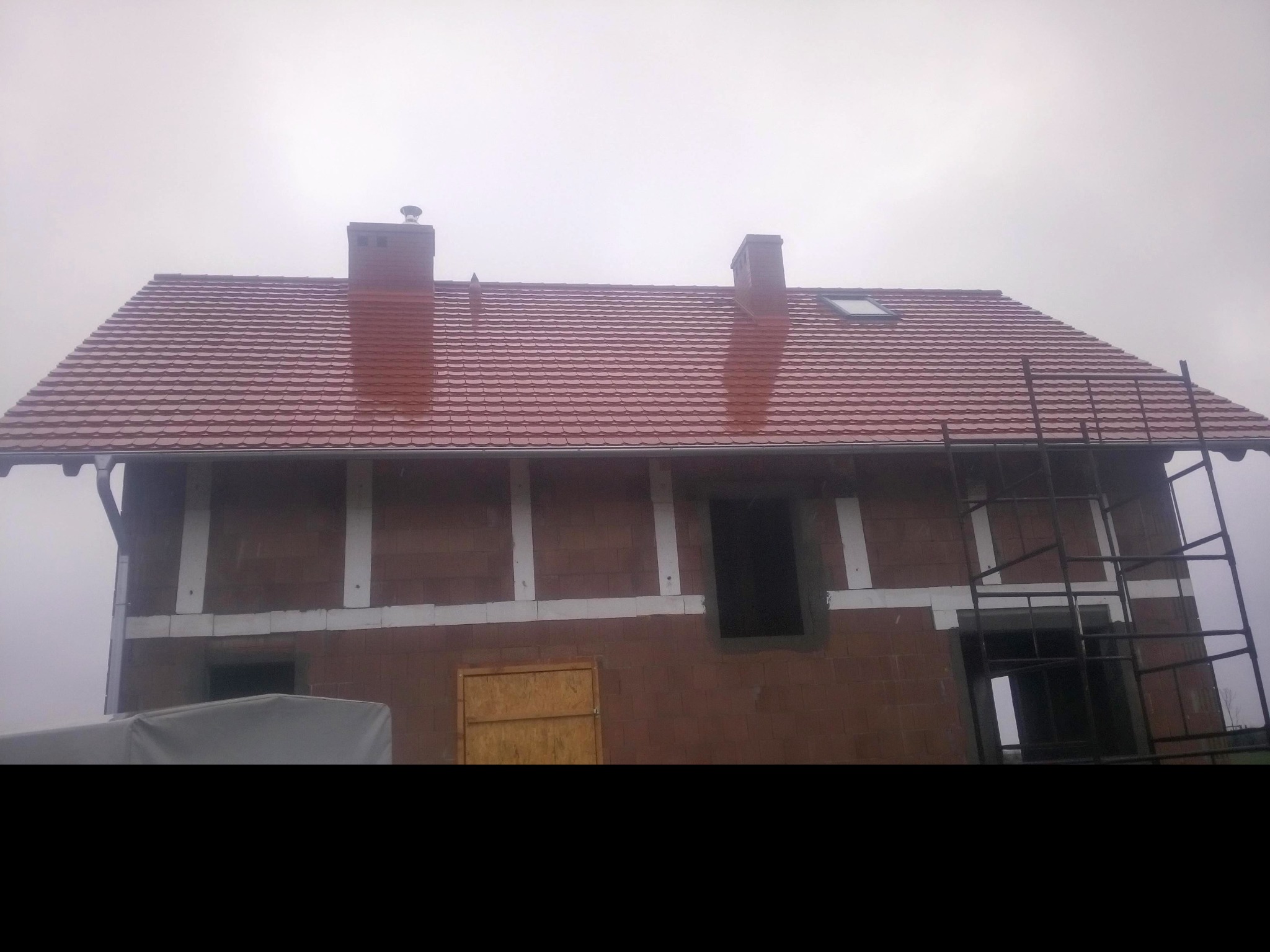 Czerwony dach z dachówki ceramicznej na budynku w trakcie budowy, widoczne kominy i okno dachowe. Na ścianach budynku widoczne białe elementy konstrukcyjne.