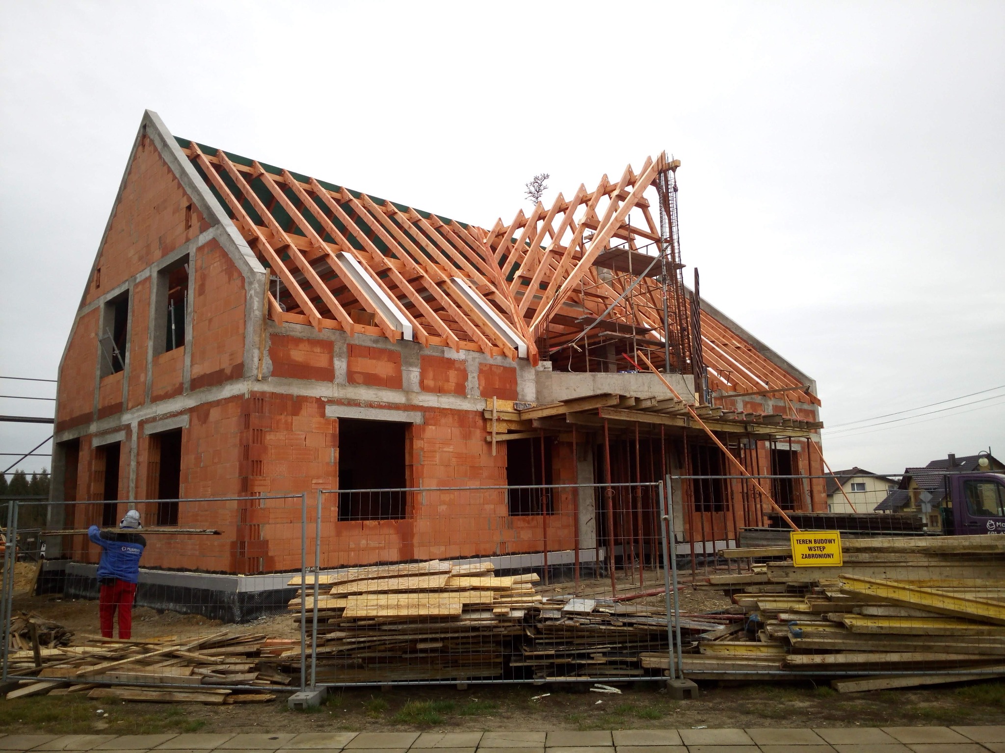 Budowa domu z czerwonej cegły: widoczna konstrukcja dachu z drewnianych belek, ogrodzenie placu budowy i tablica informacyjna. W tle pochmurne niebo.