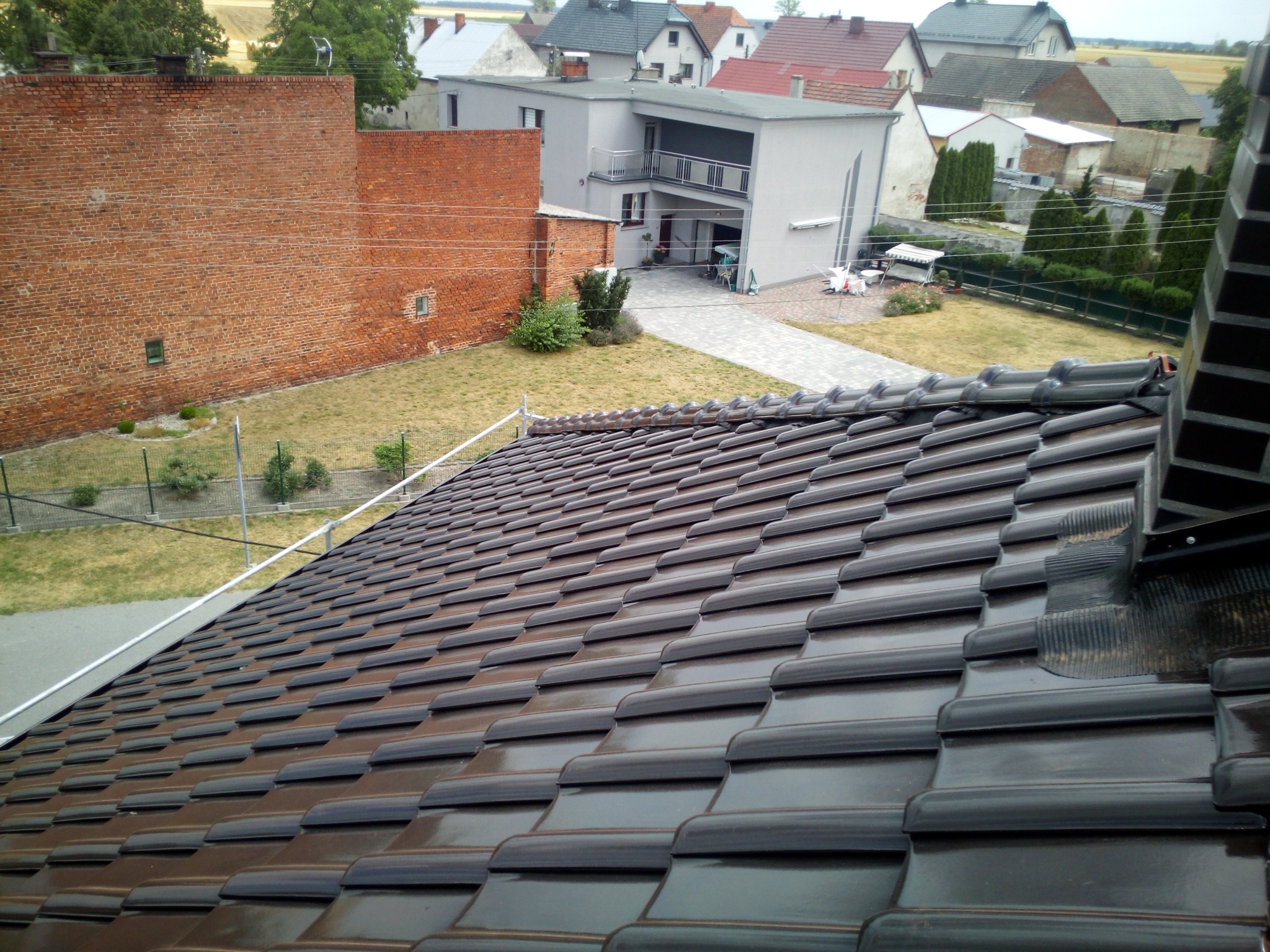 Nowy, błyszczący dach z ciemnej dachówki ceramicznej, widok z góry na tle zabudowań i zieleni. Układ dachówek w regularnych rzędach, z widocznymi detalami łączeń.