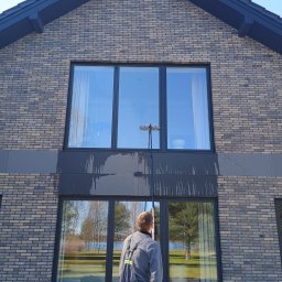 DEMINU USŁUGI MYJĄCE - Pracownik w kombinezonie myje okna na wysokości w nowoczesnym, dwupiętrowym domu z cegły. Widoczne smugi brudnej wody na elewacji. Używa teleskopowego kija z mopem.