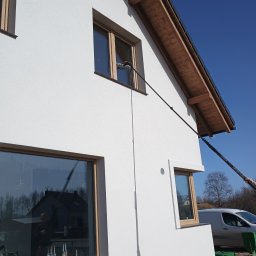 DEMINU USŁUGI MYJĄCE - Biała elewacja domu z drewnianą stolarką okienną, czyszczona teleskopową lancą z poziomu gruntu. Widoczny fragment dachu i błękitne niebo.