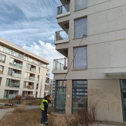 DEMINU USŁUGI MYJĄCE - Pracownik w kamizelce odblaskowej czyści okna na wysokości w nowoczesnym budynku mieszkalnym za pomocą specjalistycznego sprzętu teleskopowego. Widoczne chmury na niebie.