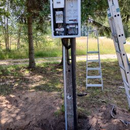 MAR VOLT - Otwarta skrzynka elektryczna z bezpiecznikami zamontowana na słupie, obok drabina. Wykopana ziemia wokół słupa sugeruje prace instalacyjne w terenie z drzewami.