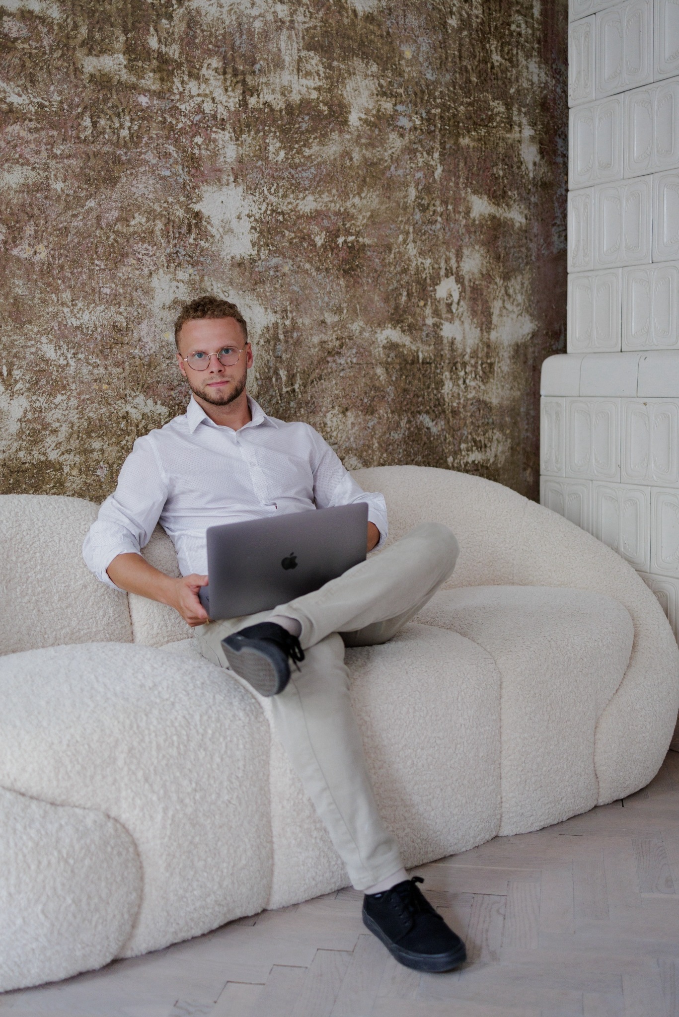 Młody mężczyzna w okularach siedzi na białej sofie z laptopem na kolanach, w tle ściana o fakturze betonu i biały piec kaflowy. Ubrany w białą koszulę i beżowe spodnie.