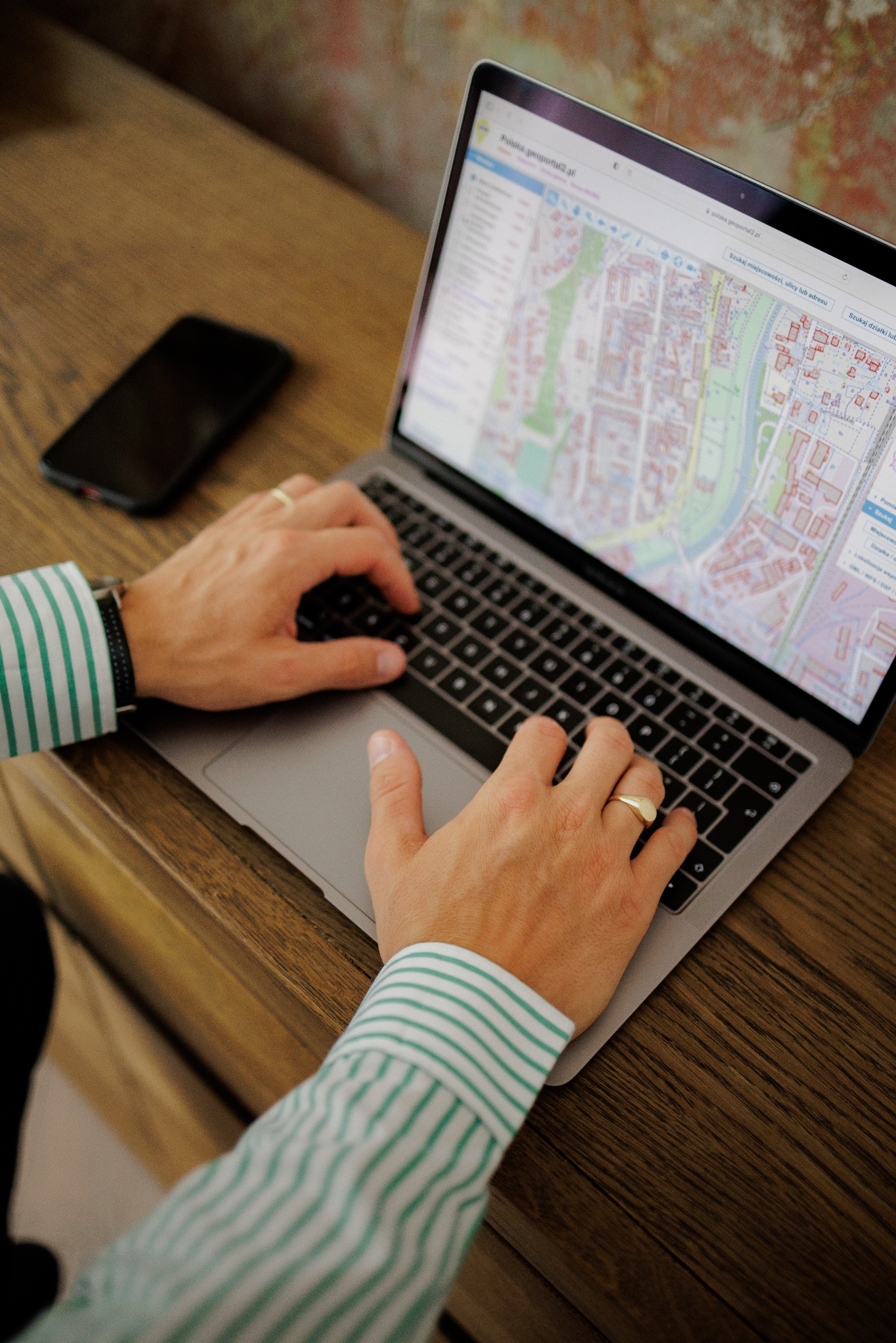 Osoba wpisuje dane na laptopie z widoczną mapą geodezyjną. Na drewnianym biurku leży smartfon. Ujęcie z góry, skupione na dłoniach i ekranie komputera.