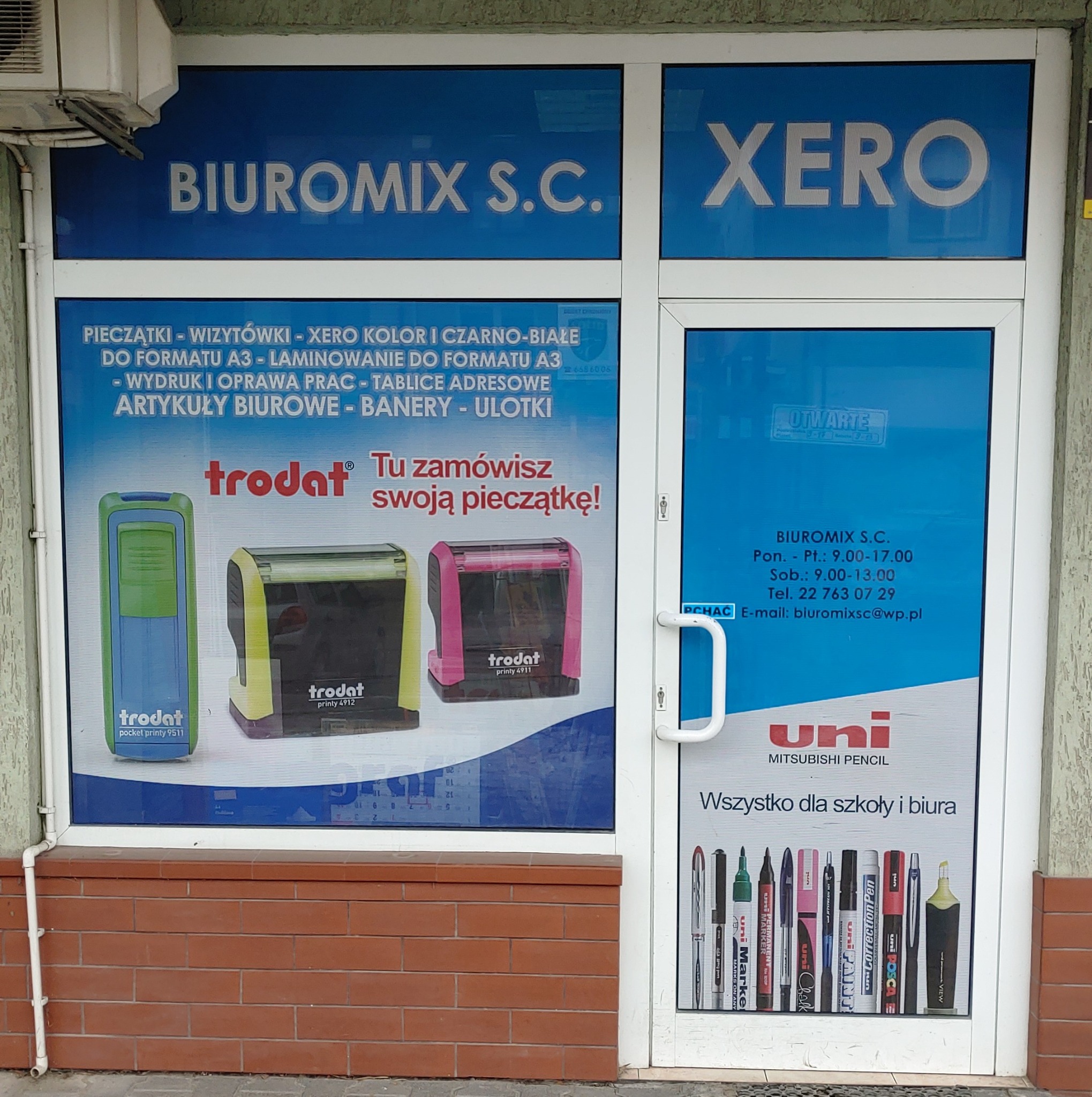 Szyld firmy Biuromix S.C. z ofertą: pieczątki, ksero, artykuły biurowe, banery, ulotki. Na szyldzie widoczne pieczątki Trodat i artykuły biurowe Uni.