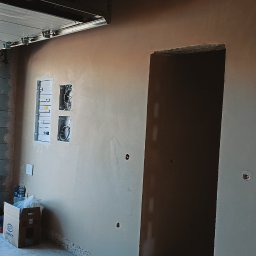 Pro-Tynki - Ściana po nałożeniu gładzi gipsowej, widoczne puszki elektryczne i otwór drzwiowy. W tle otwarty garaż z widokiem na zewnątrz. Jasne, naturalne światło.