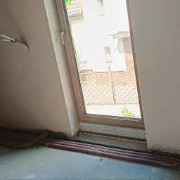 Pro-Tynki - Ściana po nałożeniu tynku gipsowego, widoczne instalacje elektryczne i rury grzewcze przy podłodze. Okno z widokiem na zewnątrz. Etap prac wykończeniowych.