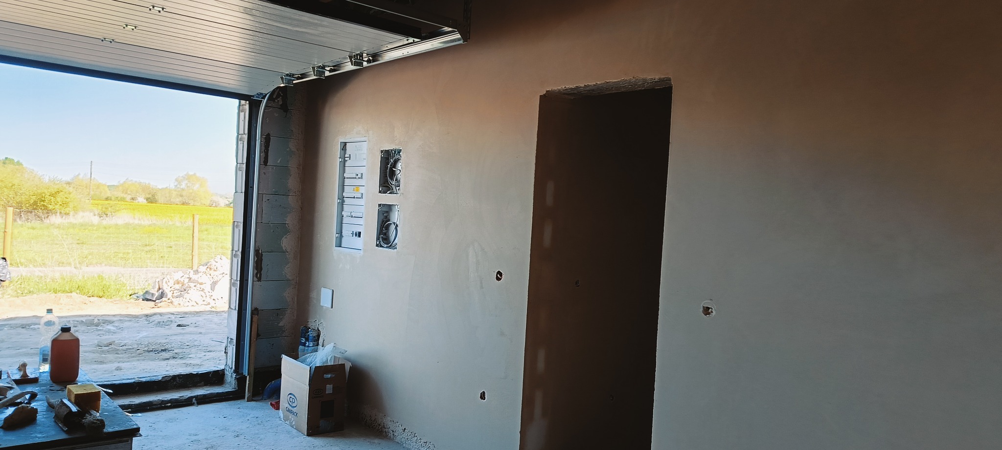 Ściana po nałożeniu gładzi gipsowej, widoczne puszki elektryczne i otwór drzwiowy. W tle otwarty garaż z widokiem na zewnątrz. Jasne, naturalne światło.