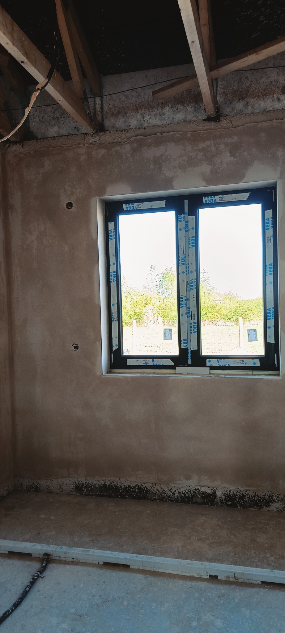 Ściana po nałożeniu tynku w surowym wnętrzu z oknem. Widoczne drewniane elementy konstrukcji dachu i instalacja elektryczna. Tynk o nierównej fakturze.