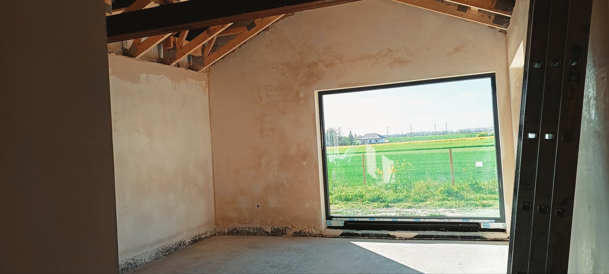 Wnętrze w trakcie prac. Ściany po nałożeniu tynku, widoczne drewniane elementy konstrukcji dachu i okno z widokiem na zielone pola. W rogu oparta drabina.