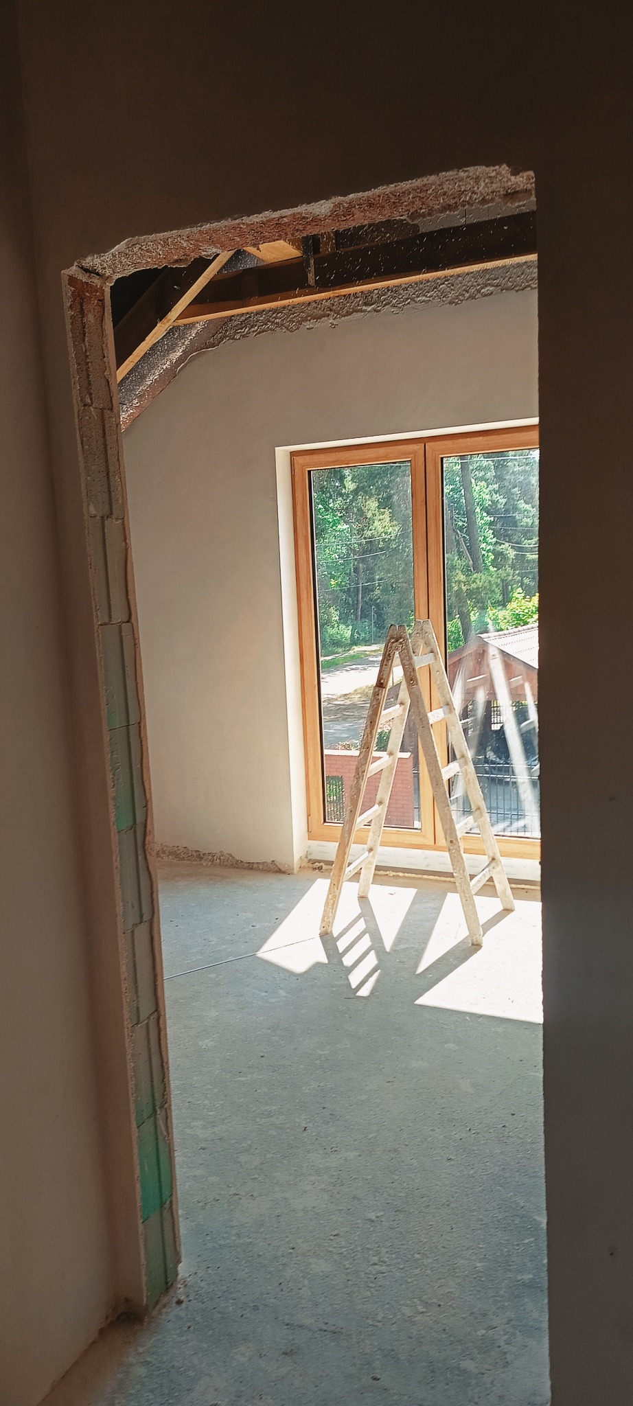 Wnętrze w trakcie prac wykończeniowych: świeżo otynkowane ściany, drewniane okno z widokiem na zieleń i drabina w jasnym świetle słonecznym.