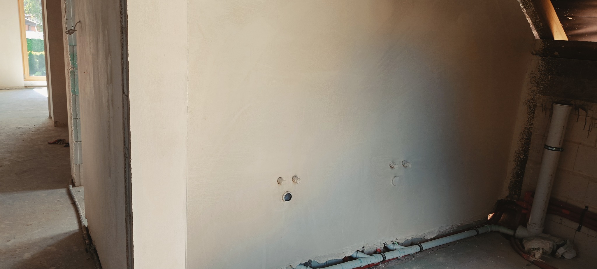 Świeżo położony, jasny tynk maszynowy na ścianie z widocznymi otworami na instalacje elektryczne i wodne. W tle widoczne surowe wnętrze budynku w trakcie wykończenia.