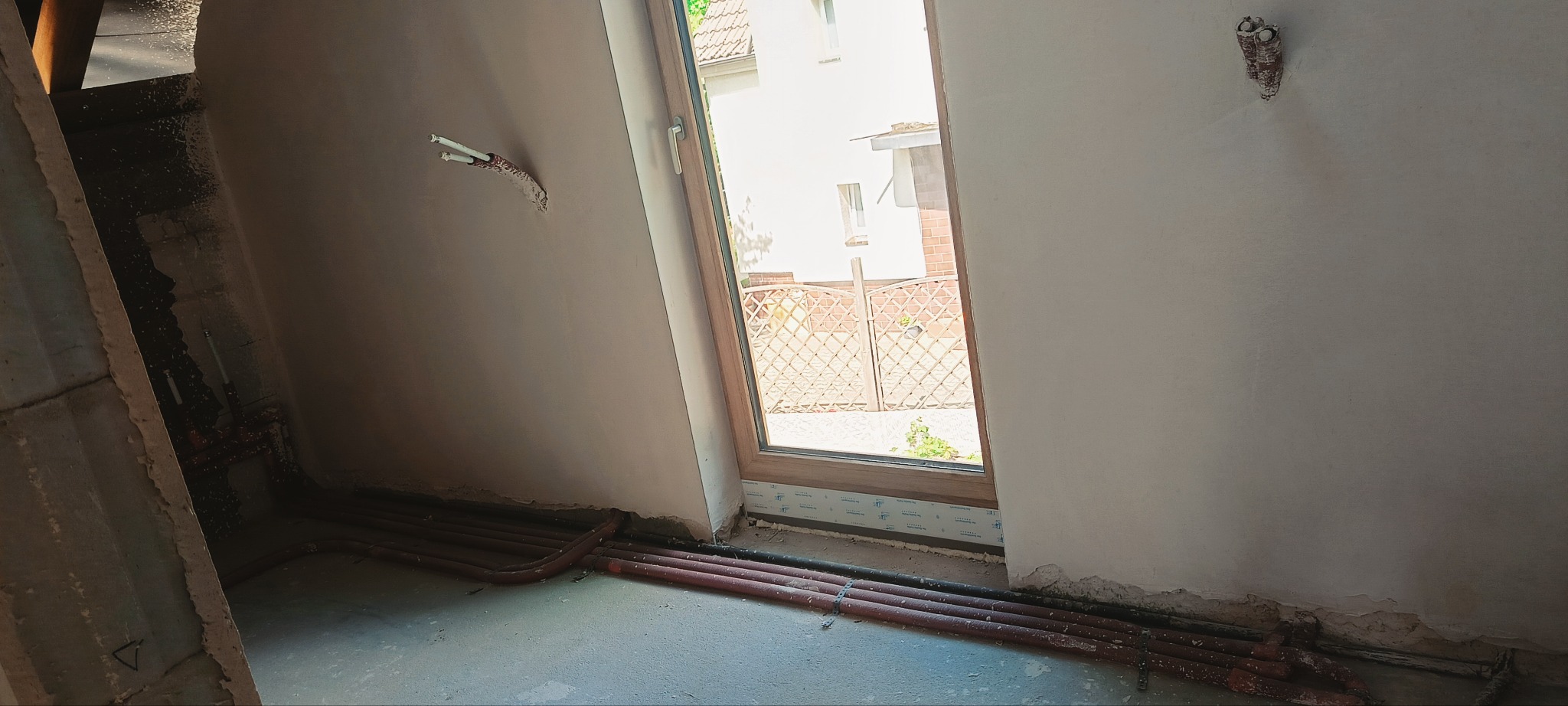 Ściana po nałożeniu tynku gipsowego, widoczne instalacje elektryczne i rury grzewcze przy podłodze. Okno z widokiem na zewnątrz. Etap prac wykończeniowych.