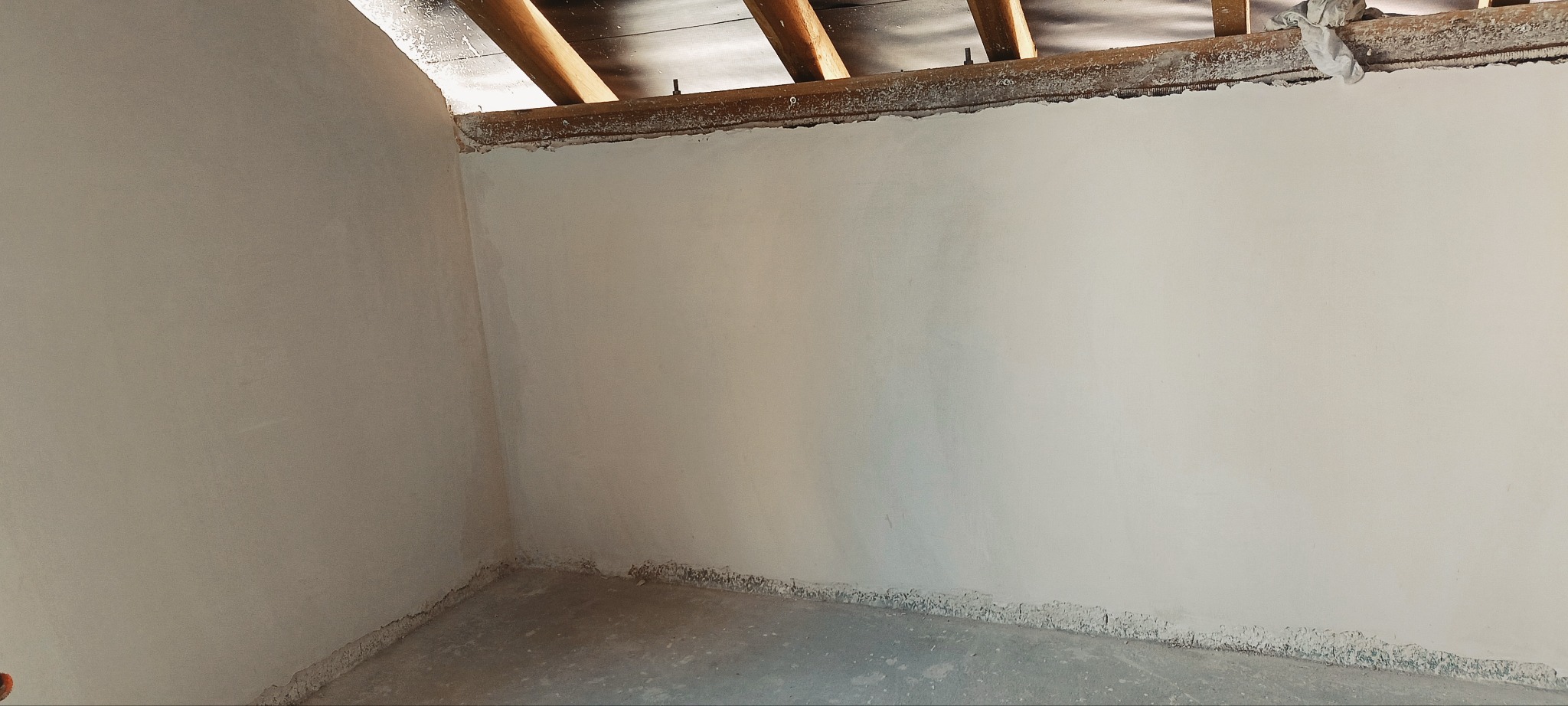 Świeżo położony, jasny tynk na ścianach wewnętrznych w rogu pomieszczenia. Widoczne drewniane elementy konstrukcyjne pod sufitem. Minimalistyczne ujęcie.