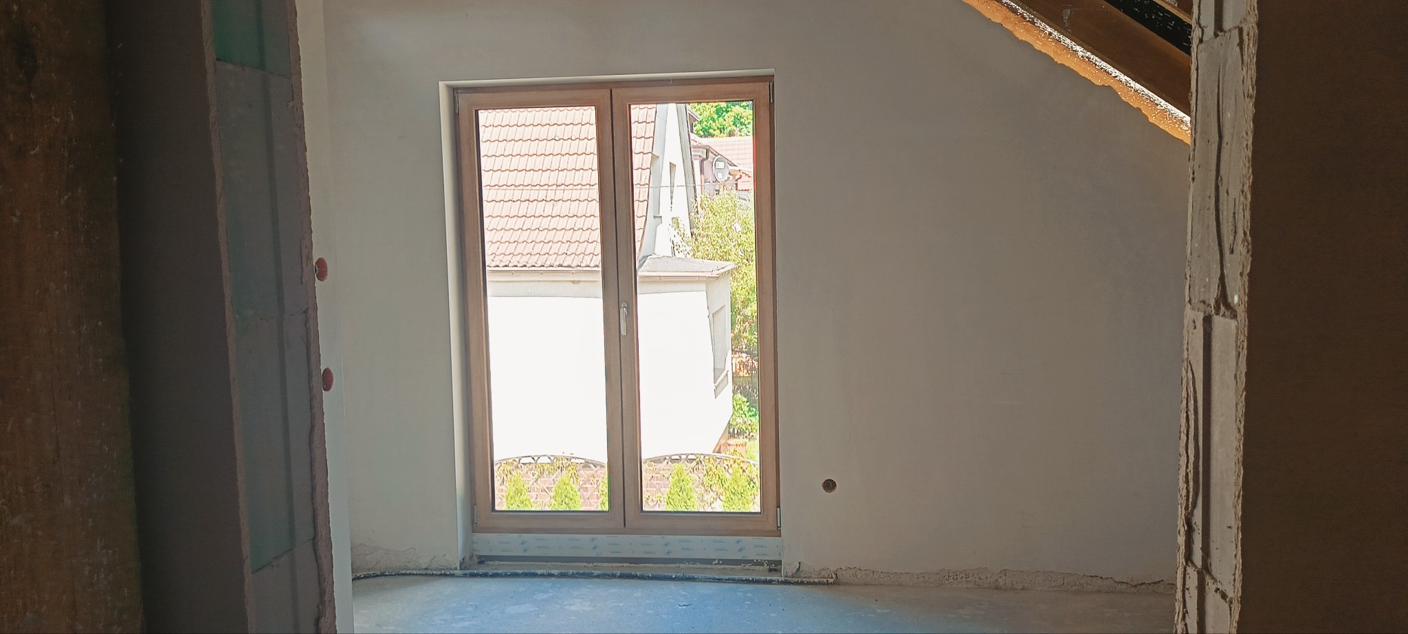 Wnętrze w trakcie tynkowania: gładkie ściany po tynkowaniu, okno z widokiem na zewnątrz, surowy stan wykończenia, widoczne ślady prac budowlanych.