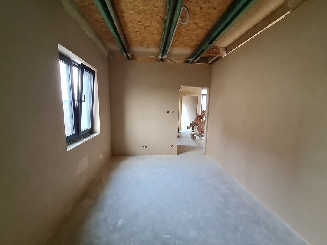 Wnętrze po nałożeniu gładzi: świeżo otynkowane ściany w jasnym odcieniu beżu, widoczne okno z czarną ramą i surowy strop z drewnianych belek. W tle widoczny sprzęt do nakładania tynków.