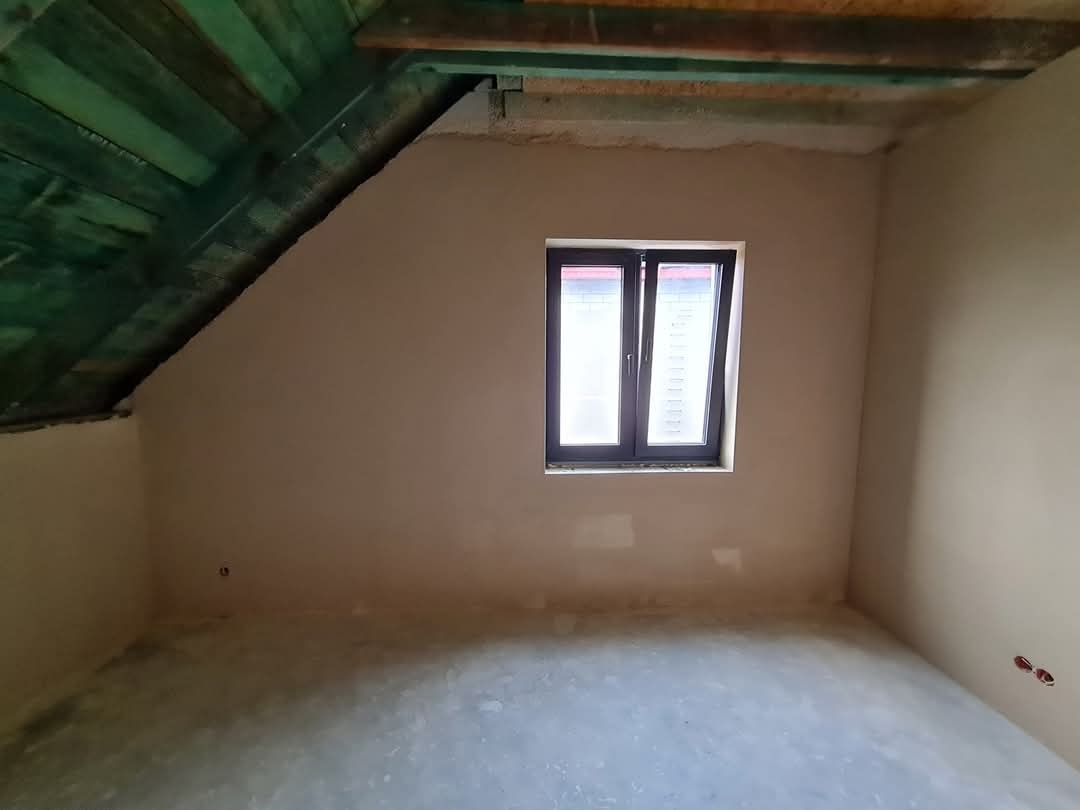 Poddasze po nałożeniu gładzi gipsowej, widoczne okno w ciemnej ramie i fragment drewnianej konstrukcji dachu pomalowanej na zielono. Ściany w kolorze beżowym.