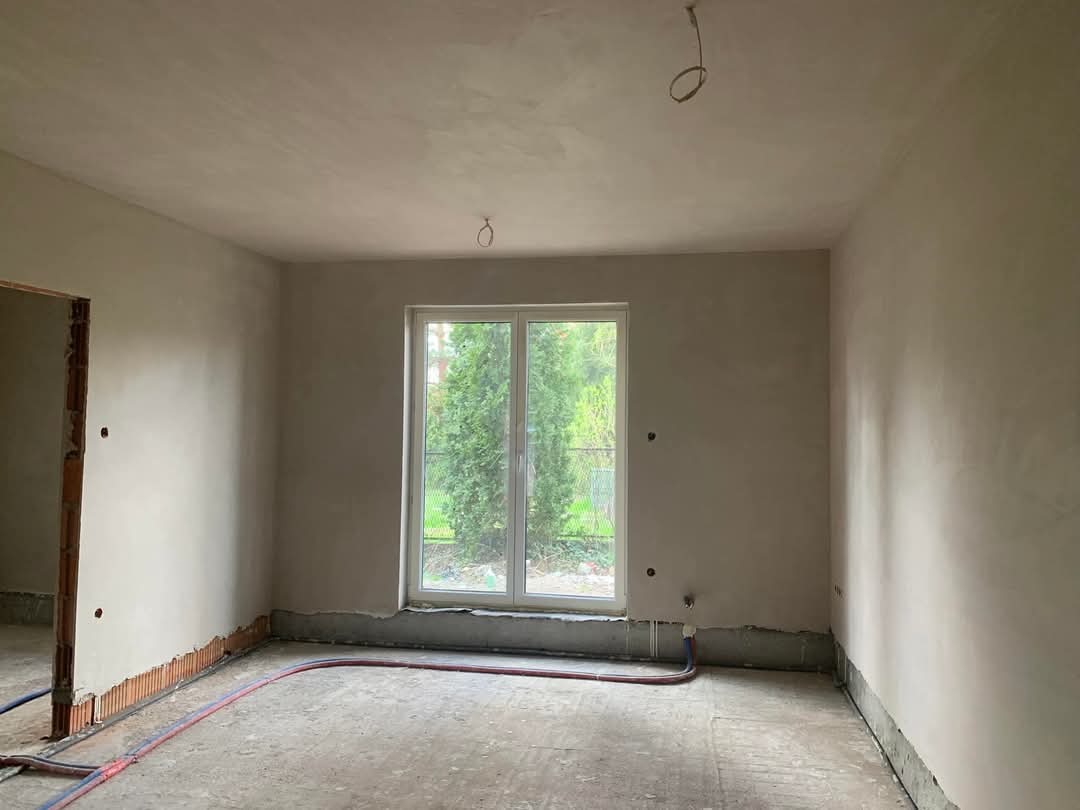 Pokój po nałożeniu gładzi gipsowej, widoczne instalacje podłogowe i okno z widokiem na zieleń. Świeżo otynkowane ściany gotowe do dalszych prac wykończeniowych.