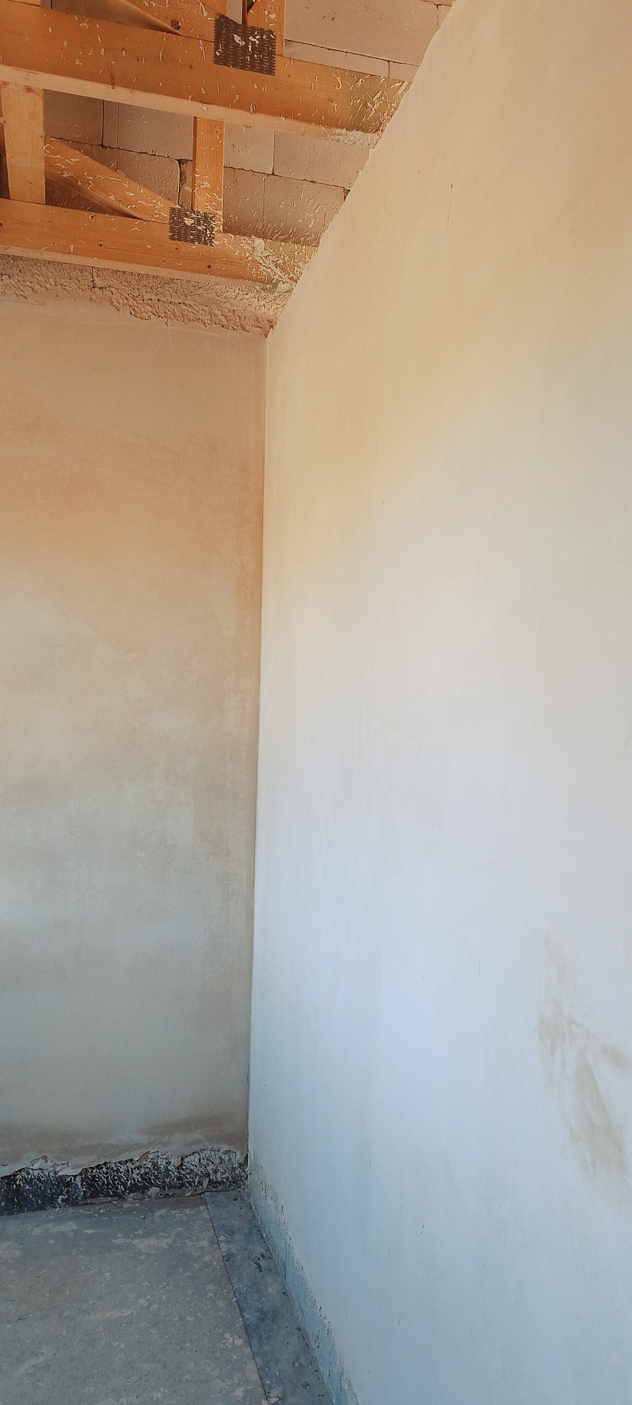 Ściana po nałożeniu tynku w surowym wnętrzu. Widoczne drewniane elementy konstrukcyjne stropu i fragment podłogi. Tynk w odcieniach beżu i bieli.