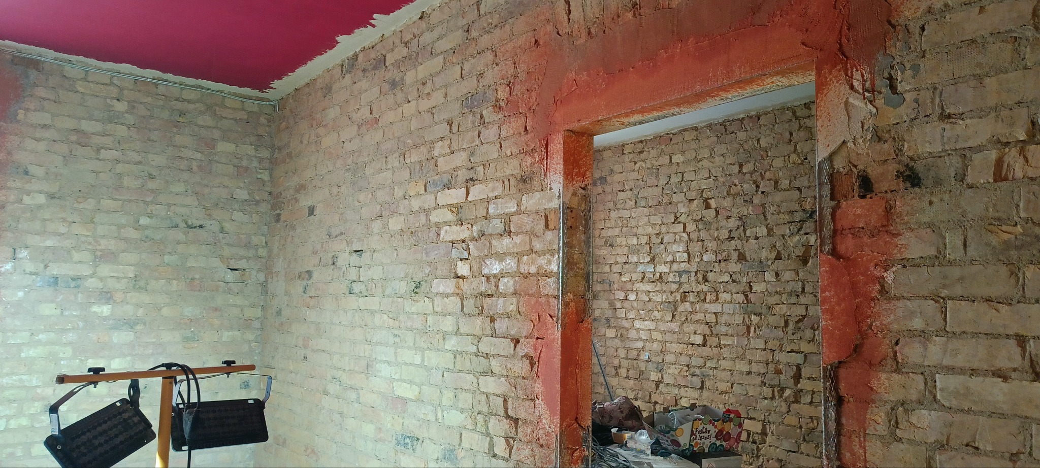 Ściana z cegły przed tynkowaniem w Łoźnicy, widoczne ślady po usuniętych starych tynkach i przygotowane nadproże. Czerwony sufit i lampa budowlana.