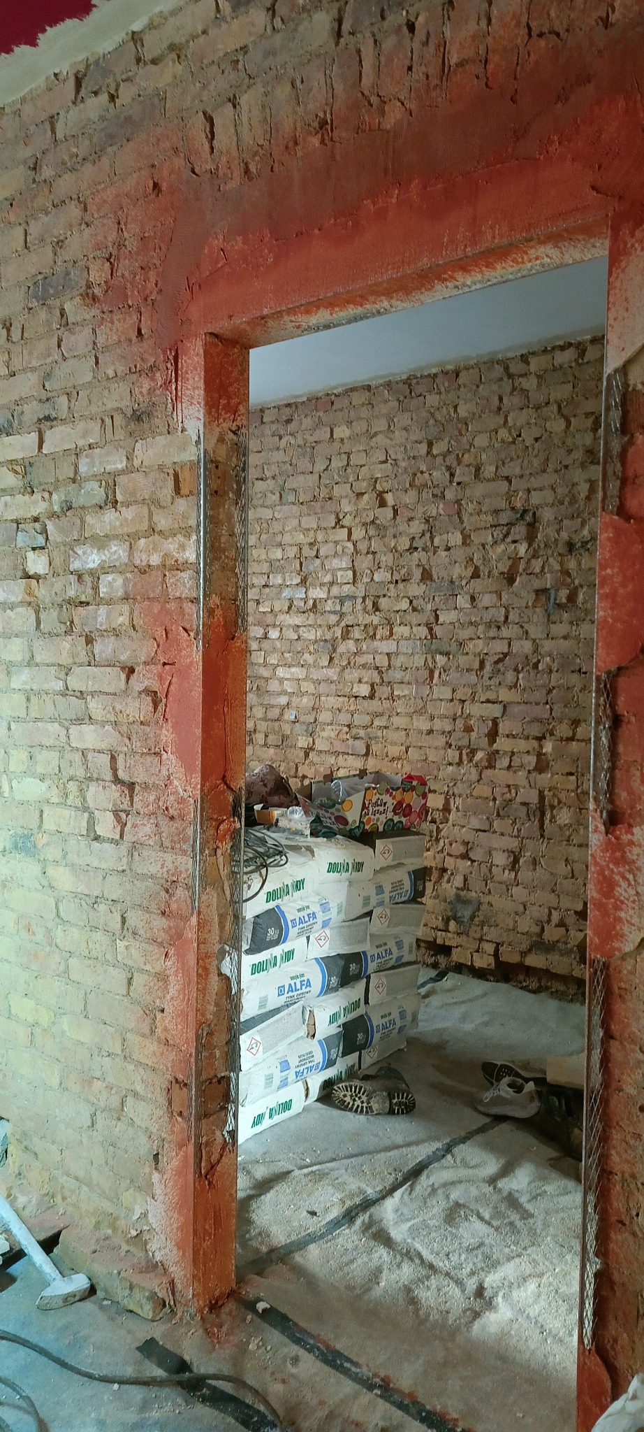Ceglana ściana w trakcie przygotowania do nałożenia tynku, widoczne worki z zaprawą Dolina Nidy Alfa i metalowa siatka na narożniku ościeżnicy.