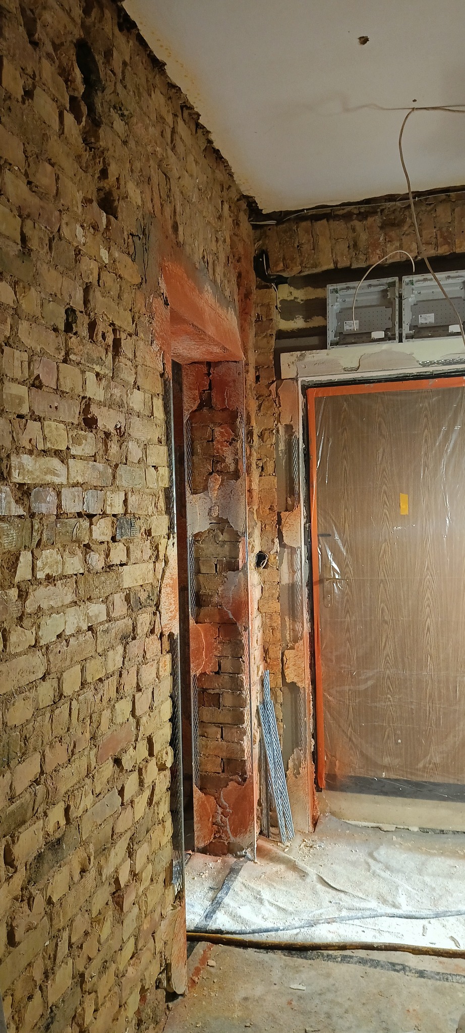 Ceglana ściana w trakcie przygotowania do tynkowania, z widocznymi siatkami wzmacniającymi wokół ościeżnicy drzwiowej. Fragment instalacji elektrycznej pod sufitem.