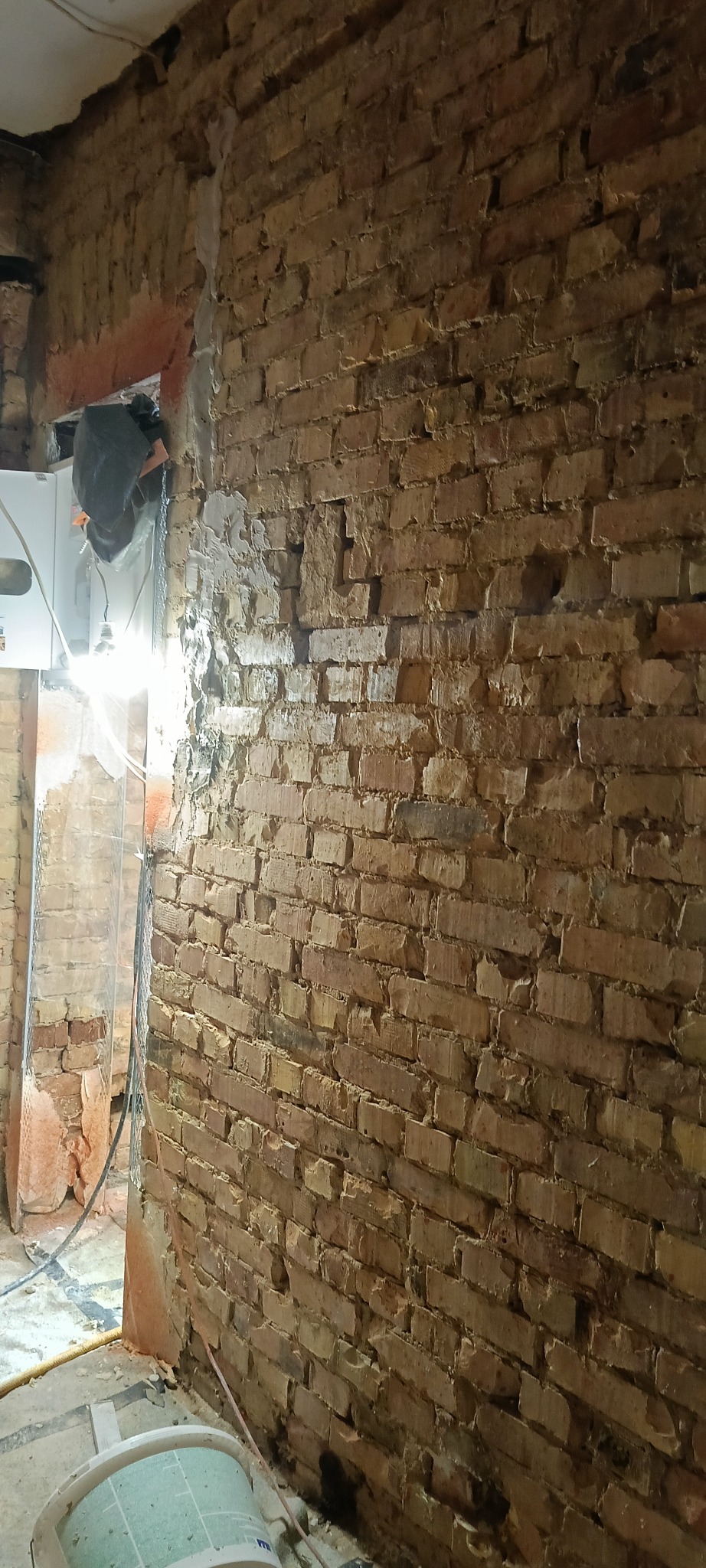 Ceglana ściana w trakcie prac przygotowawczych do tynkowania. Widoczne ślady po usuniętym starym tynku i fragment instalacji. Oświetlenie robocze.