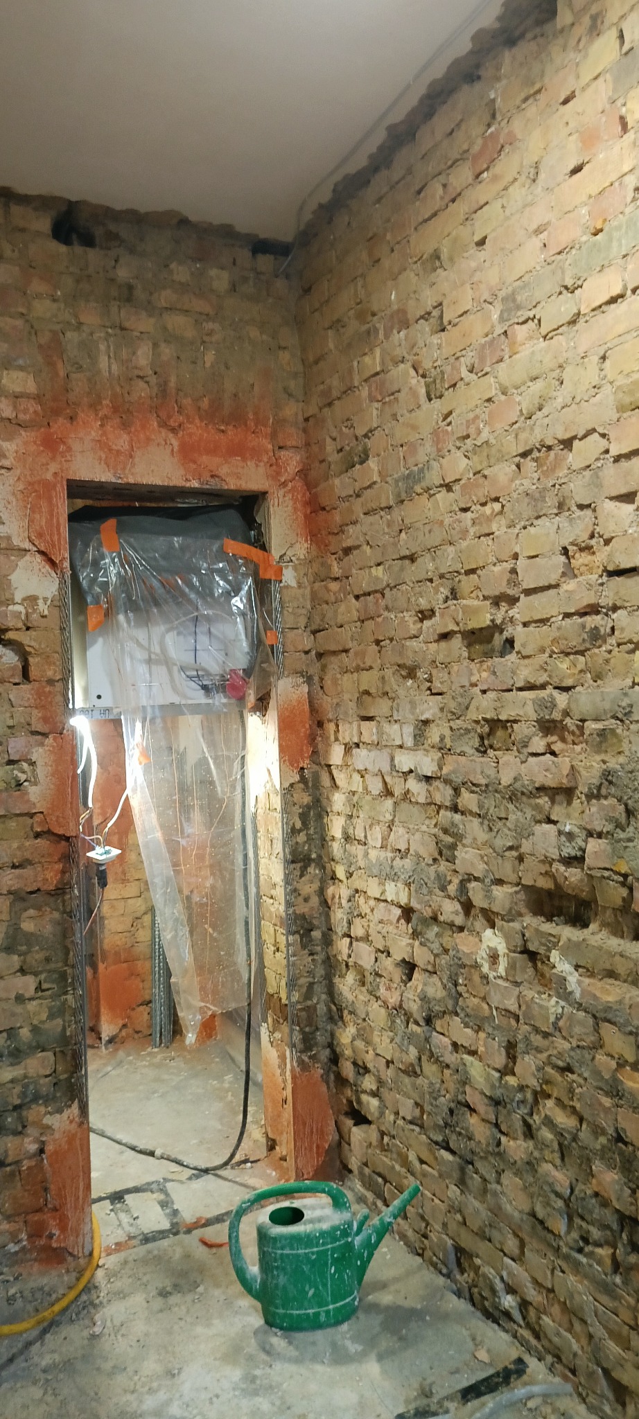 Przygotowanie do tynkowania: ściana z cegły, zabezpieczone folią drzwi, widoczna instalacja elektryczna i zielona konewka na podłodze. Surowy stan przed nałożeniem tynku.