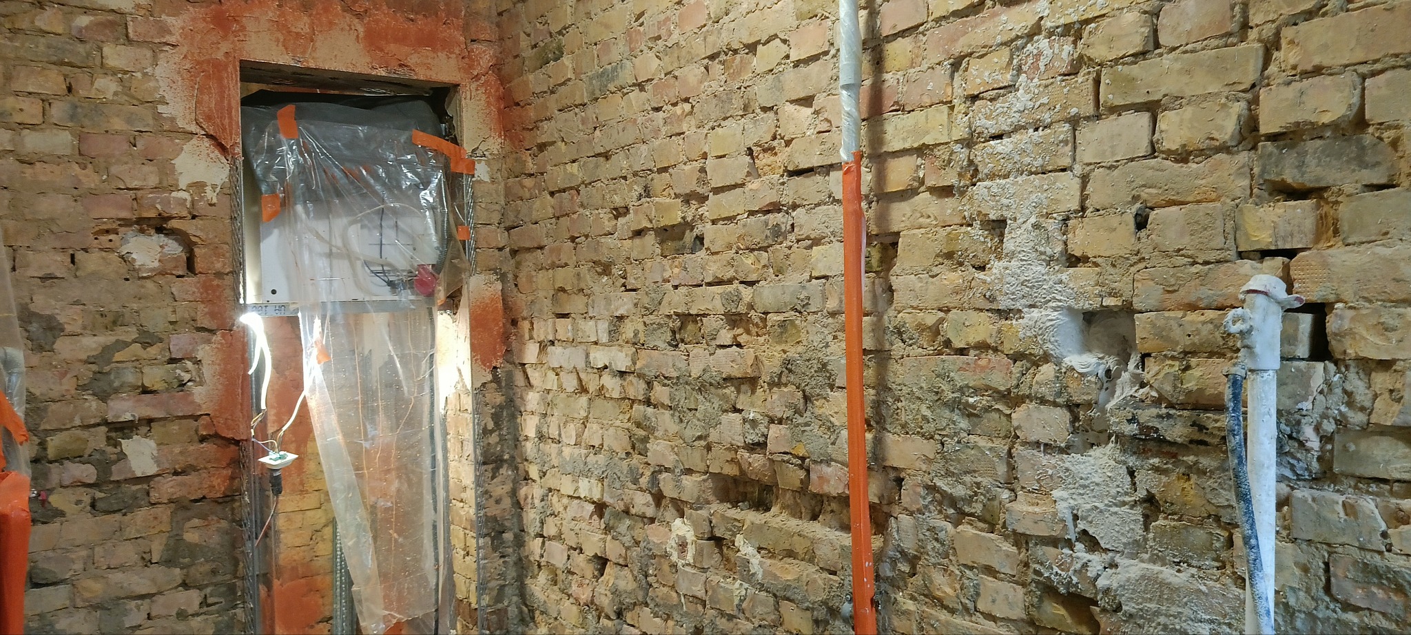 Ceglana ściana przed tynkowaniem z widocznymi instalacjami zabezpieczonymi folią i rurami. Fragmenty ściany z ubytkami i pozostałościami zaprawy.