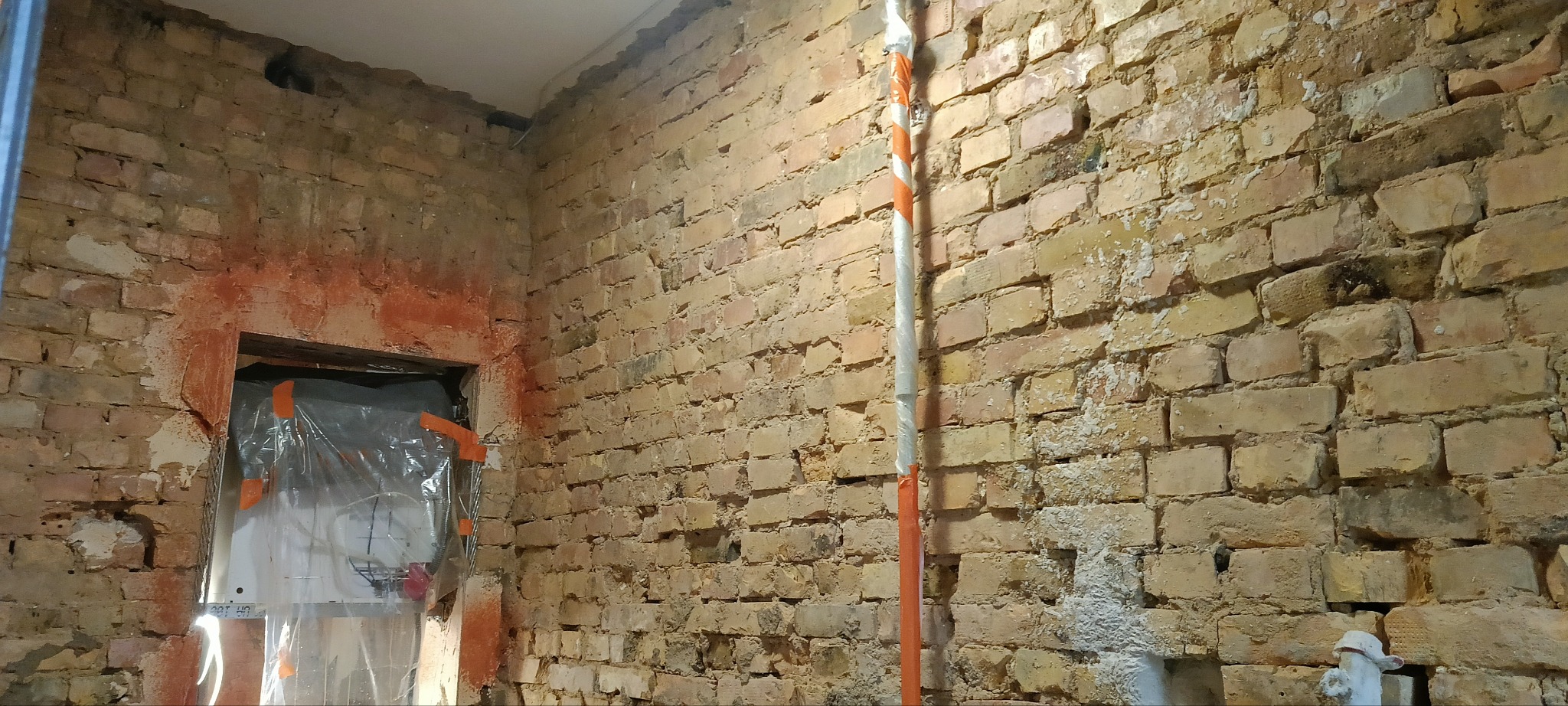 Surowa ściana z cegły przed tynkowaniem, zabezpieczone drzwi folią, widoczne ślady po instalacjach. Przygotowanie do prac wykończeniowych w pomieszczeniu.