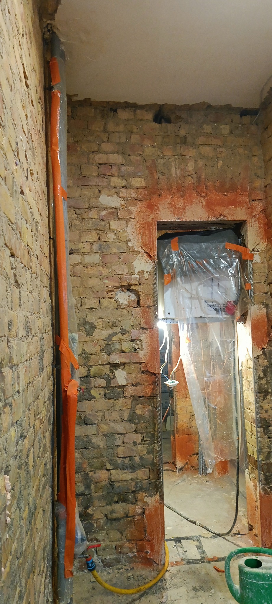 Przygotowanie do prac tynkarskich: zabezpieczone rury, folia ochronna na ścianie z cegły, widoczne ślady po tynkowaniu. W tle instalacja elektryczna.