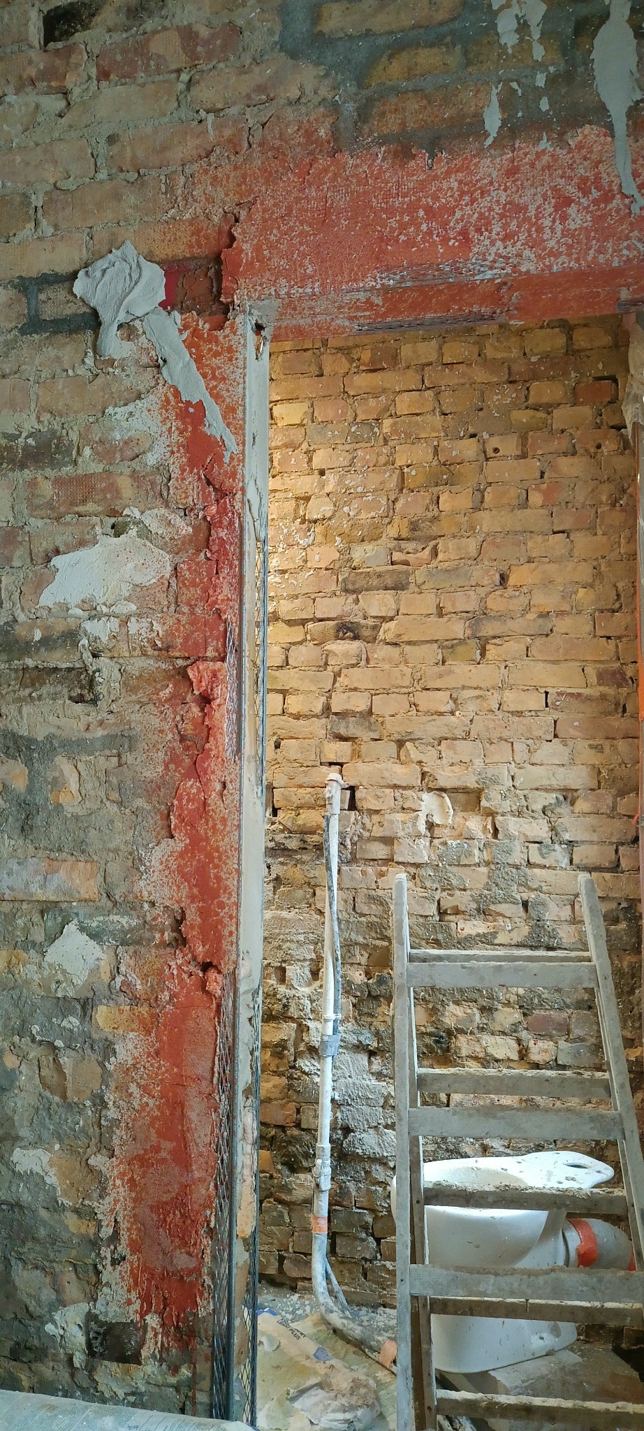 Przygotowanie ściany z cegły do nałożenia tynku: siatka, częściowo nałożona warstwa wyrównująca, widoczne instalacje i drabina. Prace remontowe w trakcie.